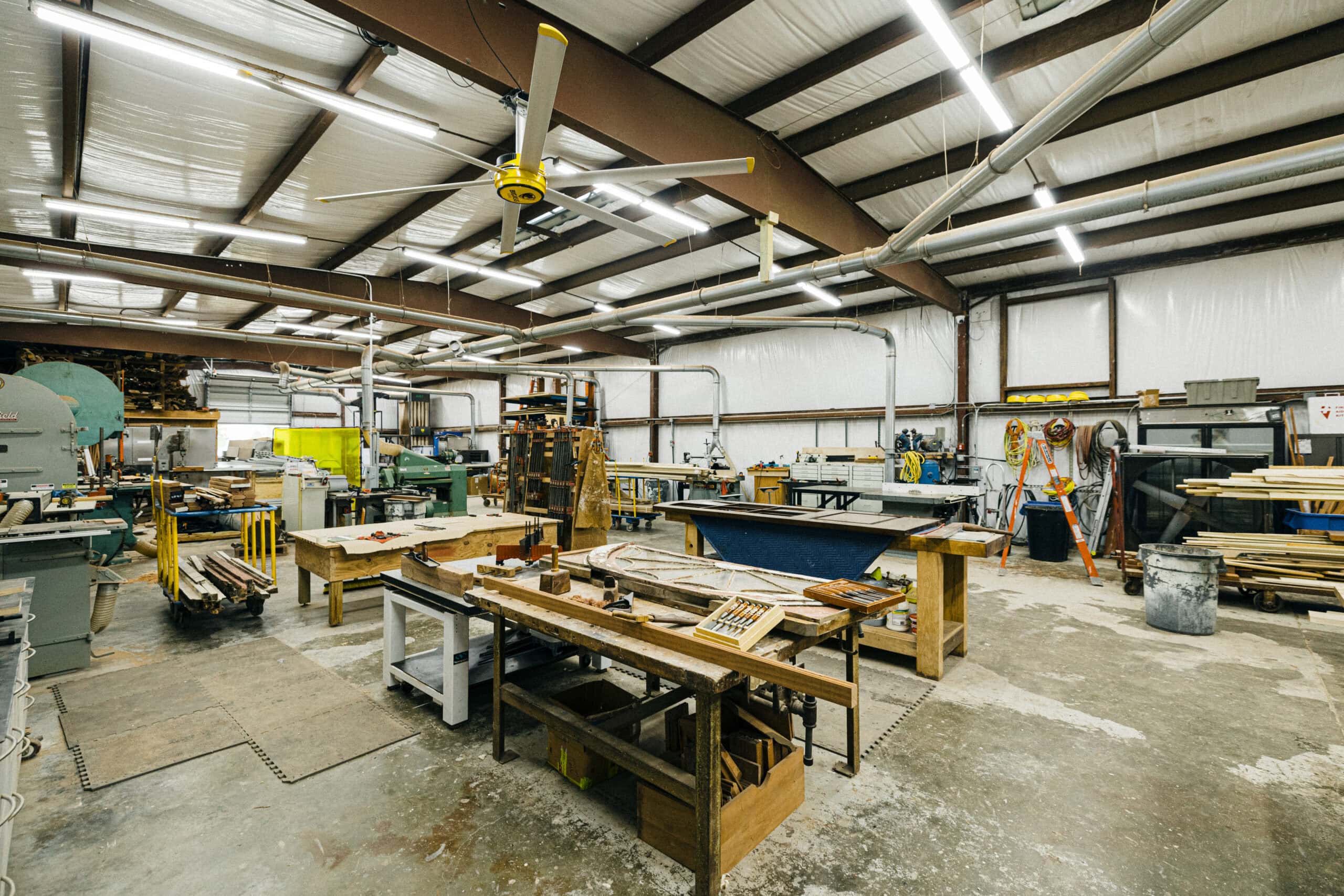 A spacious workshop with workbenches, woodworking tools, stacks of lumber, and industrial machinery. The ceiling has exposed beams, fluorescent lighting, and a large ceiling fan. Safety equipment hangs on the back wall. - McNatt Contracting