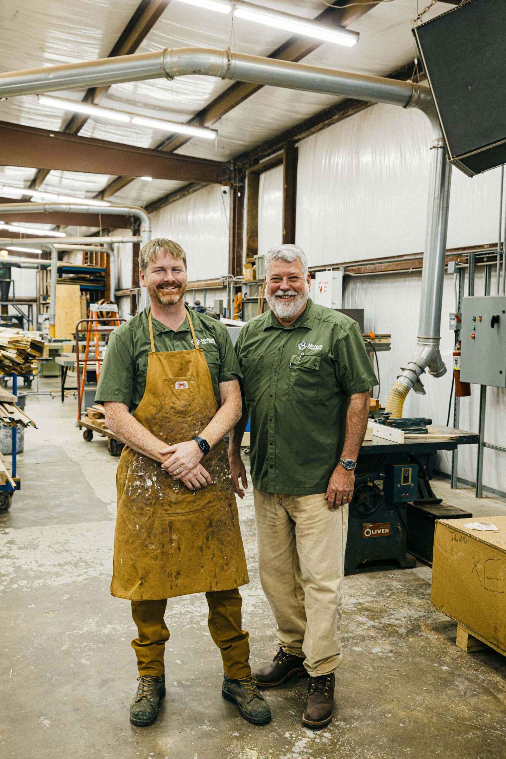 Two men stand side by side in a woodworking shop. One wears a brown apron and work clothes, the other has a green shirt and beige pants. Various woodworking tools and equipment are visible in the background. - McNatt Contracting