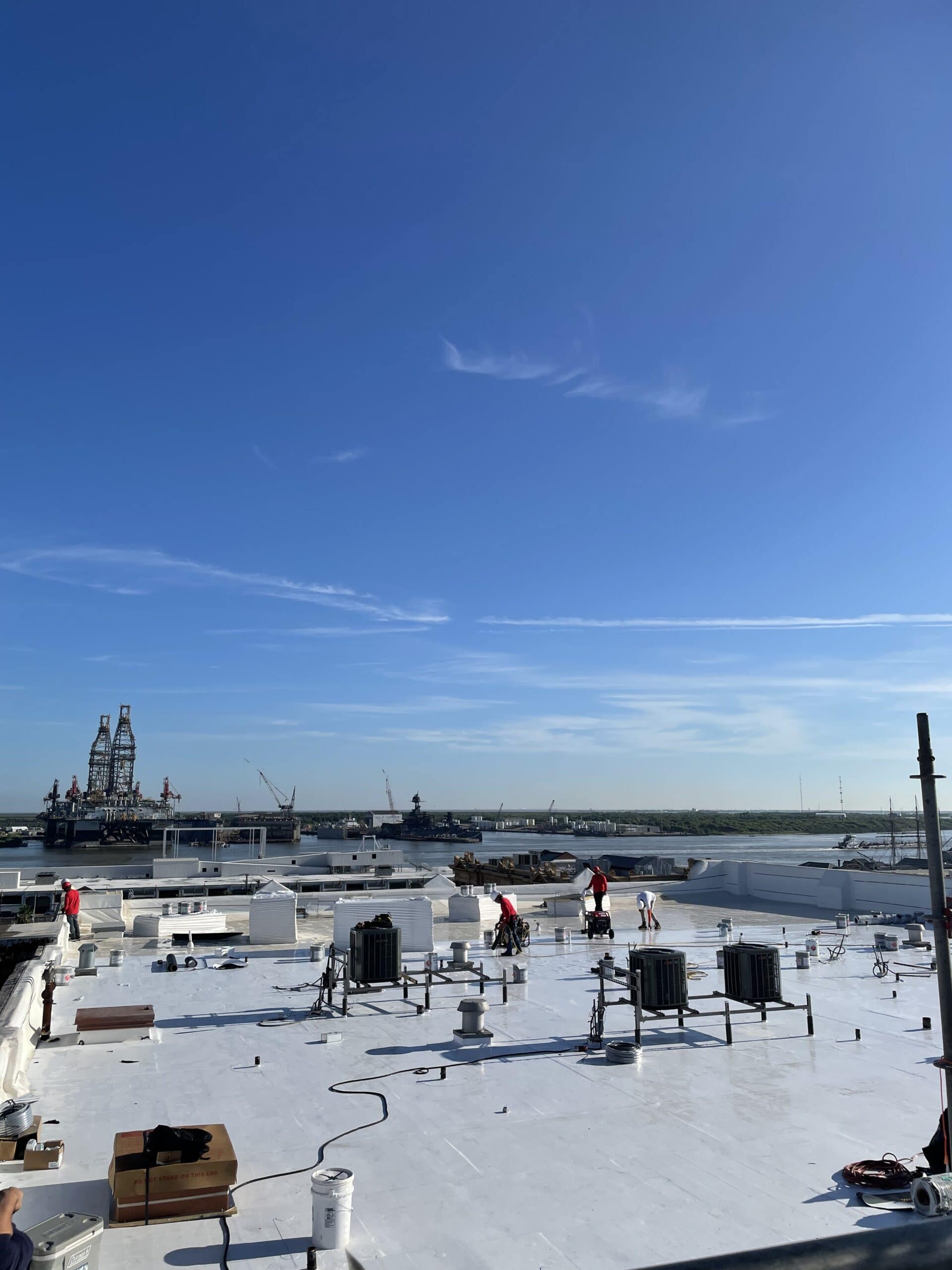Workers from McNatt Contracting are on a flat white rooftop with HVAC units and cables, under a clear blue sky. In the distance, water, cranes, ships, and industrial structures line the shore. - McNatt Contracting