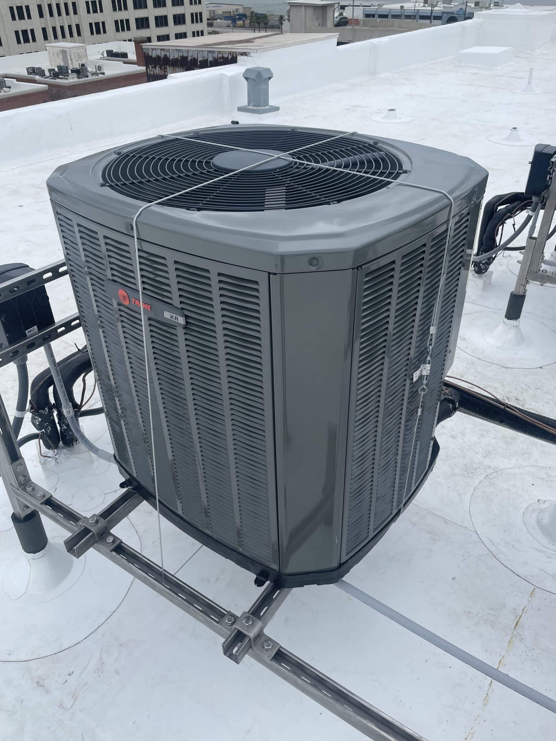 A large gray outdoor HVAC unit from McNatt Contracting is mounted on a metal platform on a flat rooftop, surrounded by utility pipes and other equipment. Nearby buildings can be seen in the background. - McNatt Contracting
