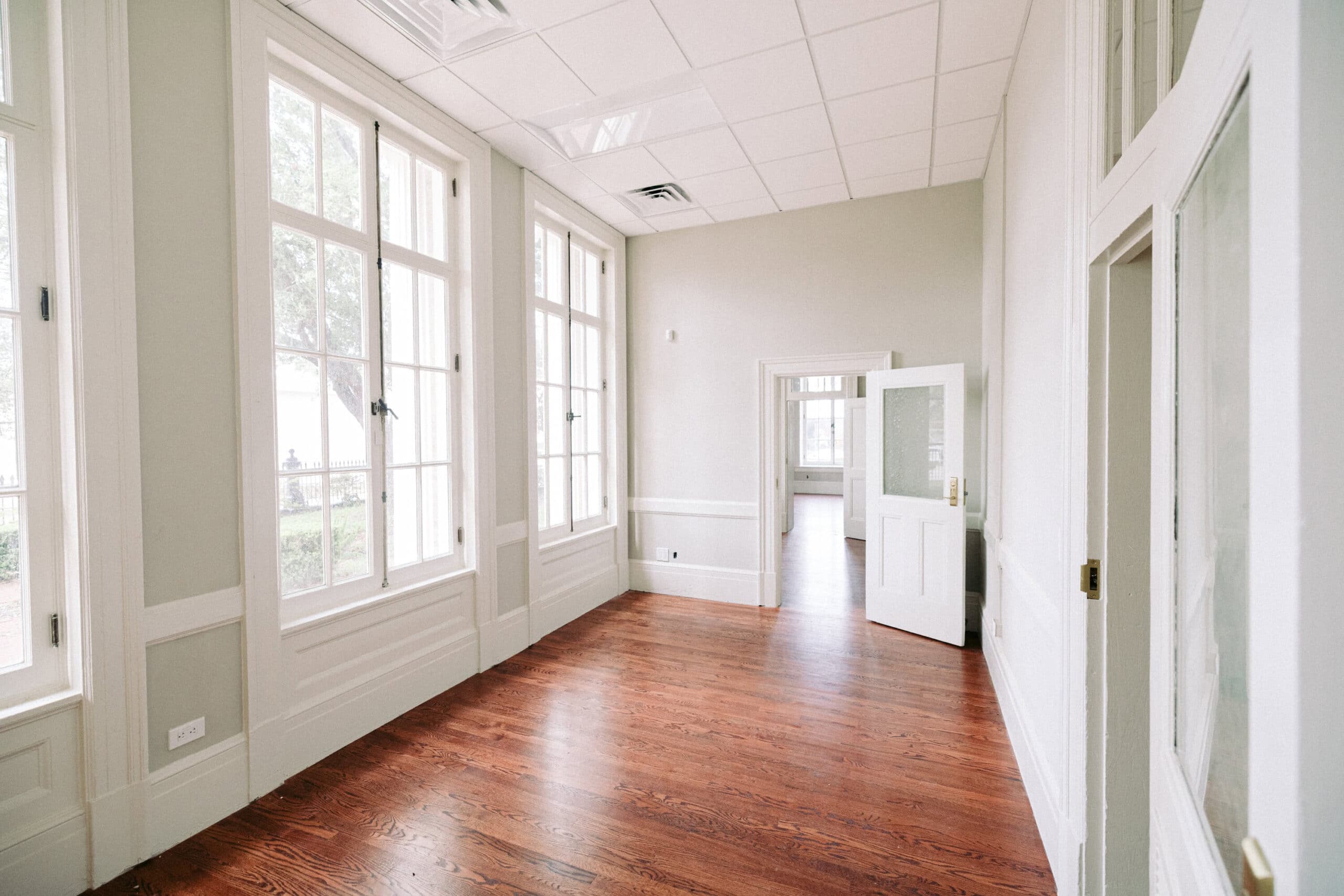 Empty room with large windows, white walls, high ceiling, and wooden floor. An open door leads to another similar room in the background. Natural light fills the space. - McNatt Contracting