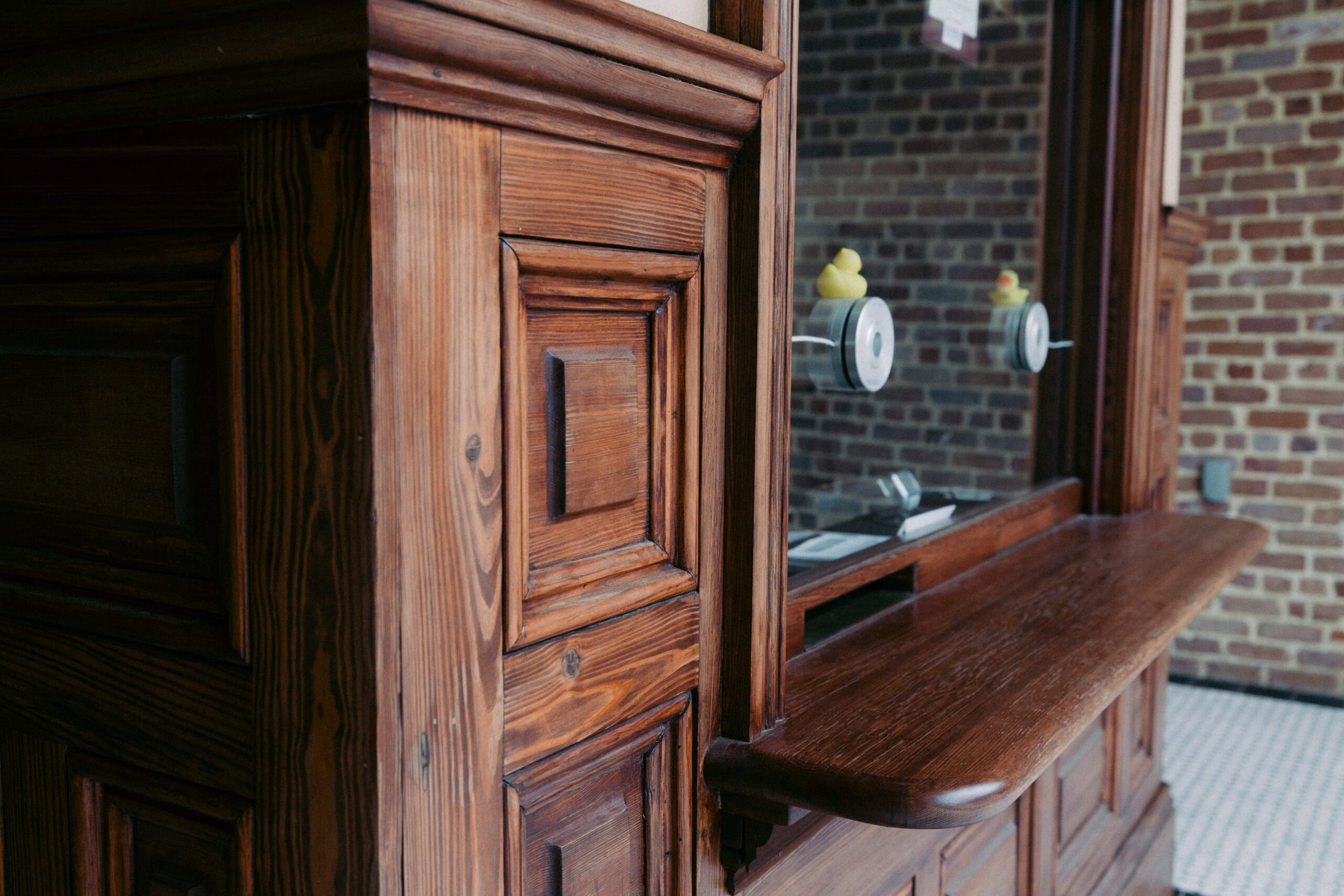 A wooden ticket counter with a glass window and two round speaker openings. A pair of rubber duck figurines sit on the ledge inside. The background features a brick wall and a tiled floor. - McNatt Contracting