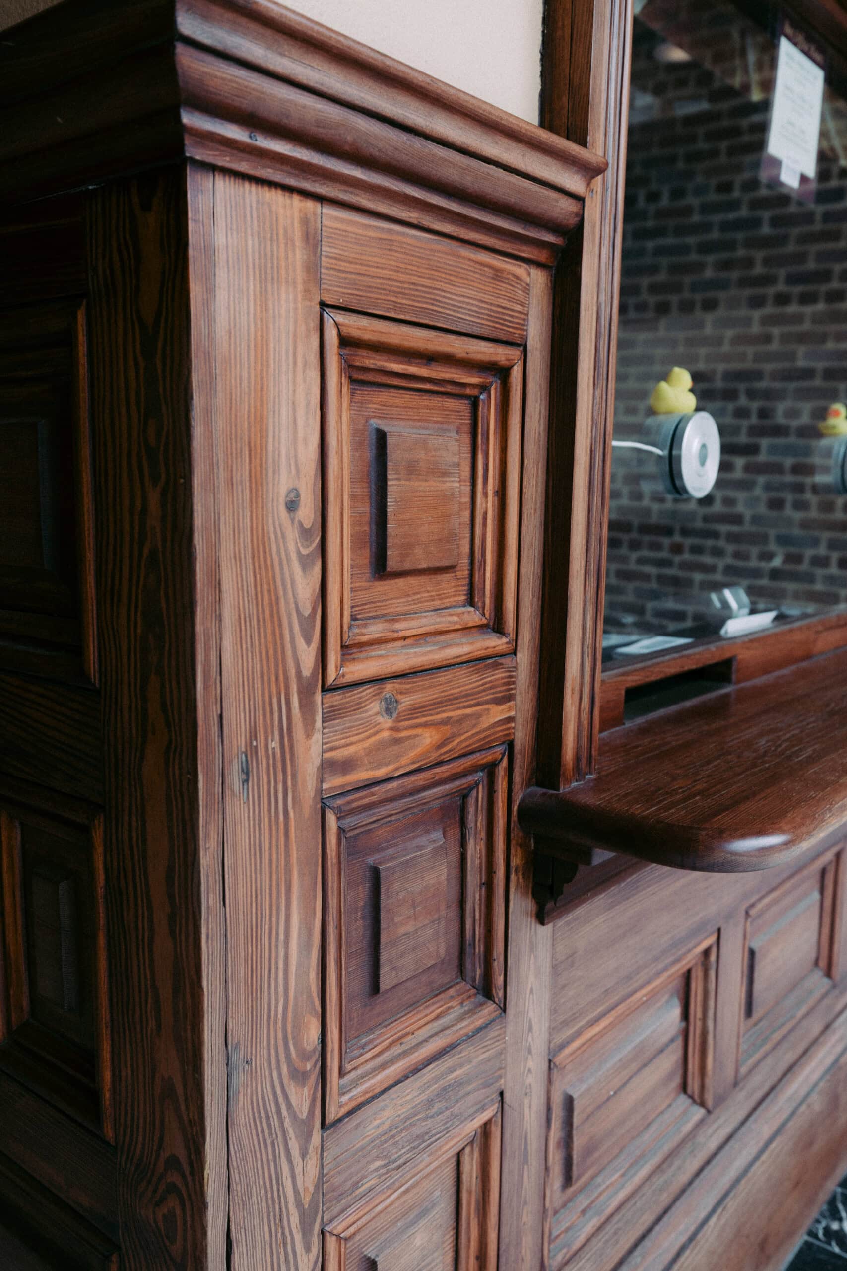 Close-up of a wooden counter and wall with decorative paneling. A glass window is set into the counter, and a brick wall is visible in the background. A small yellow rubber duck is on the window ledge. - McNatt Contracting