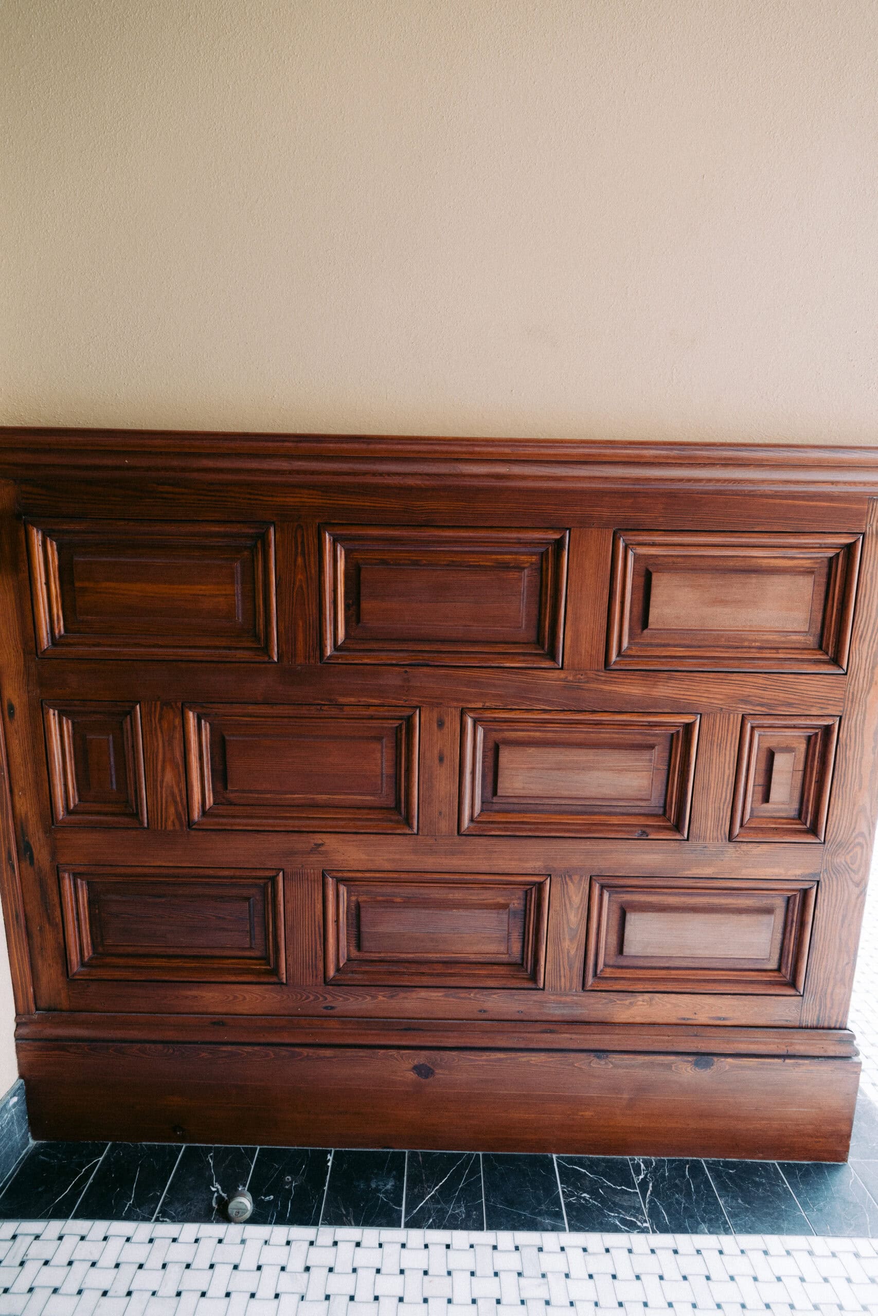 A section of wooden wall paneling with a geometric, rectangular pattern, finished in a brown stain, reminiscent of interiors at the Grand 1894 Opera House in Galveston. The wall sits above a tiled floor with a black border and white mosaic tiles. - McNatt Contracting