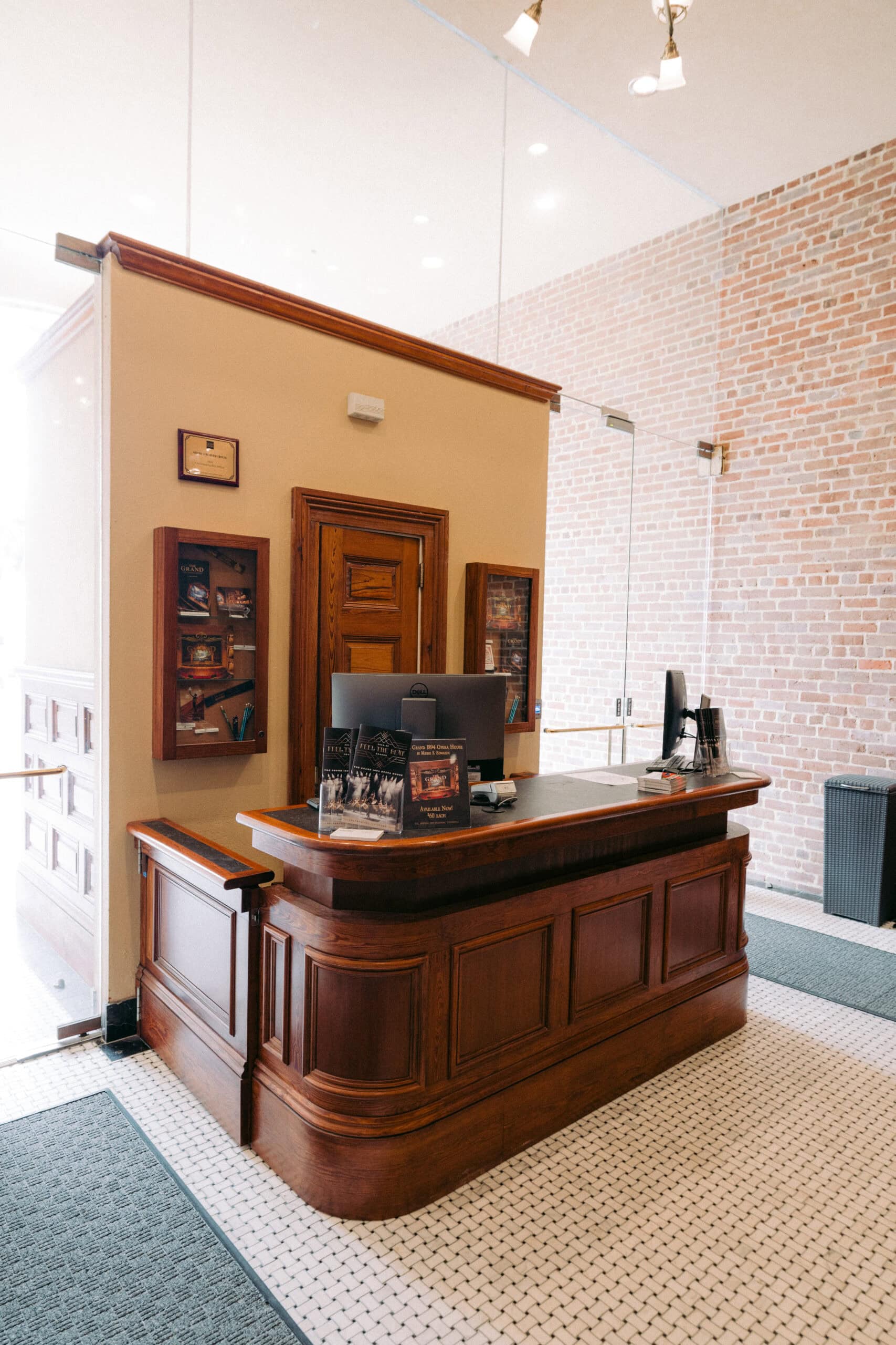 A wooden reception desk with a computer monitor, brochures, and books on top, sits in a room with brick walls, glass partitions, and tiled flooring. Shelves with items are mounted behind the desk. - McNatt Contracting