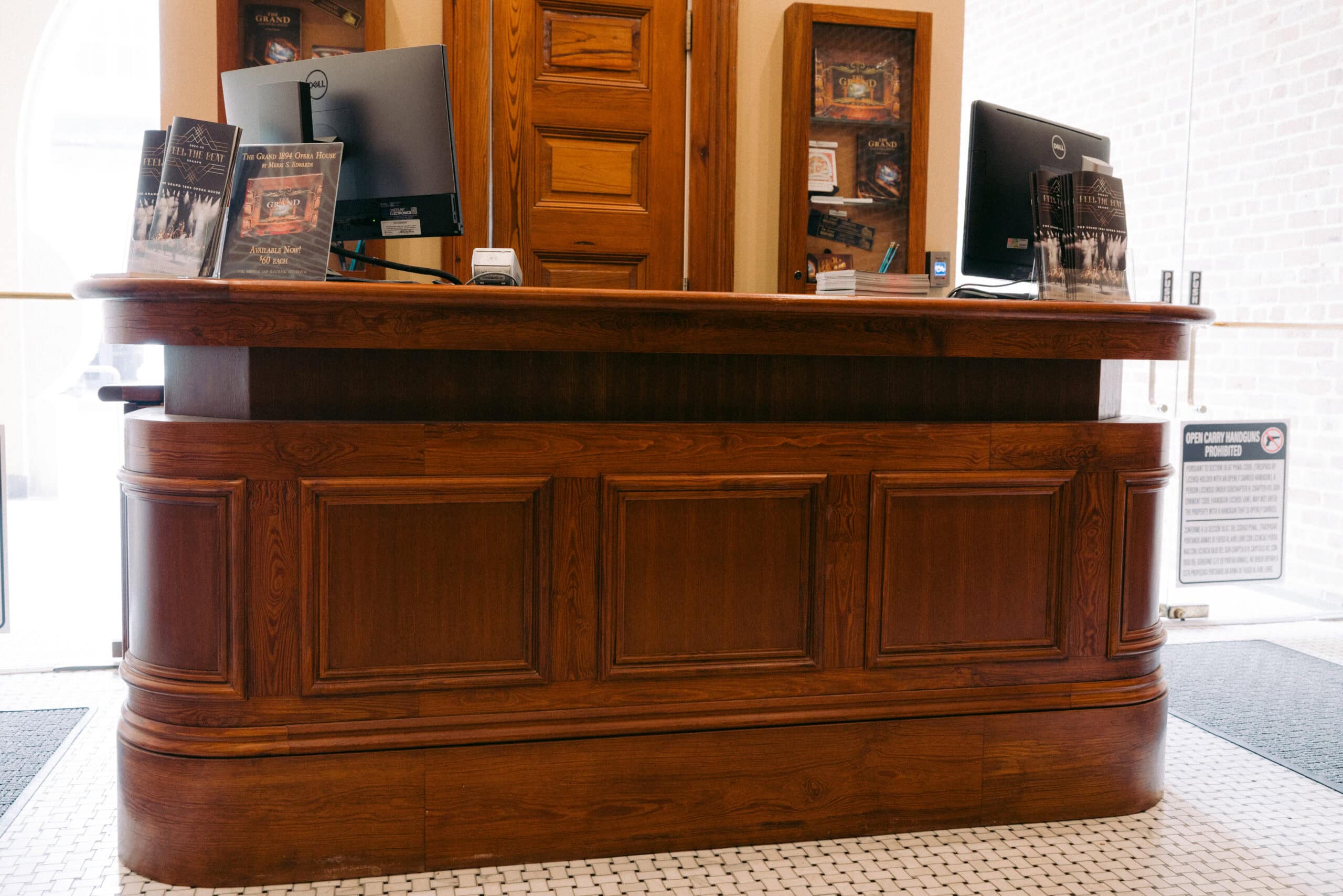 A wooden reception desk with two computer monitors, promotional brochures, and wood paneling, situated on a tiled floor with a wooden door and shelves in the background. - McNatt Contracting