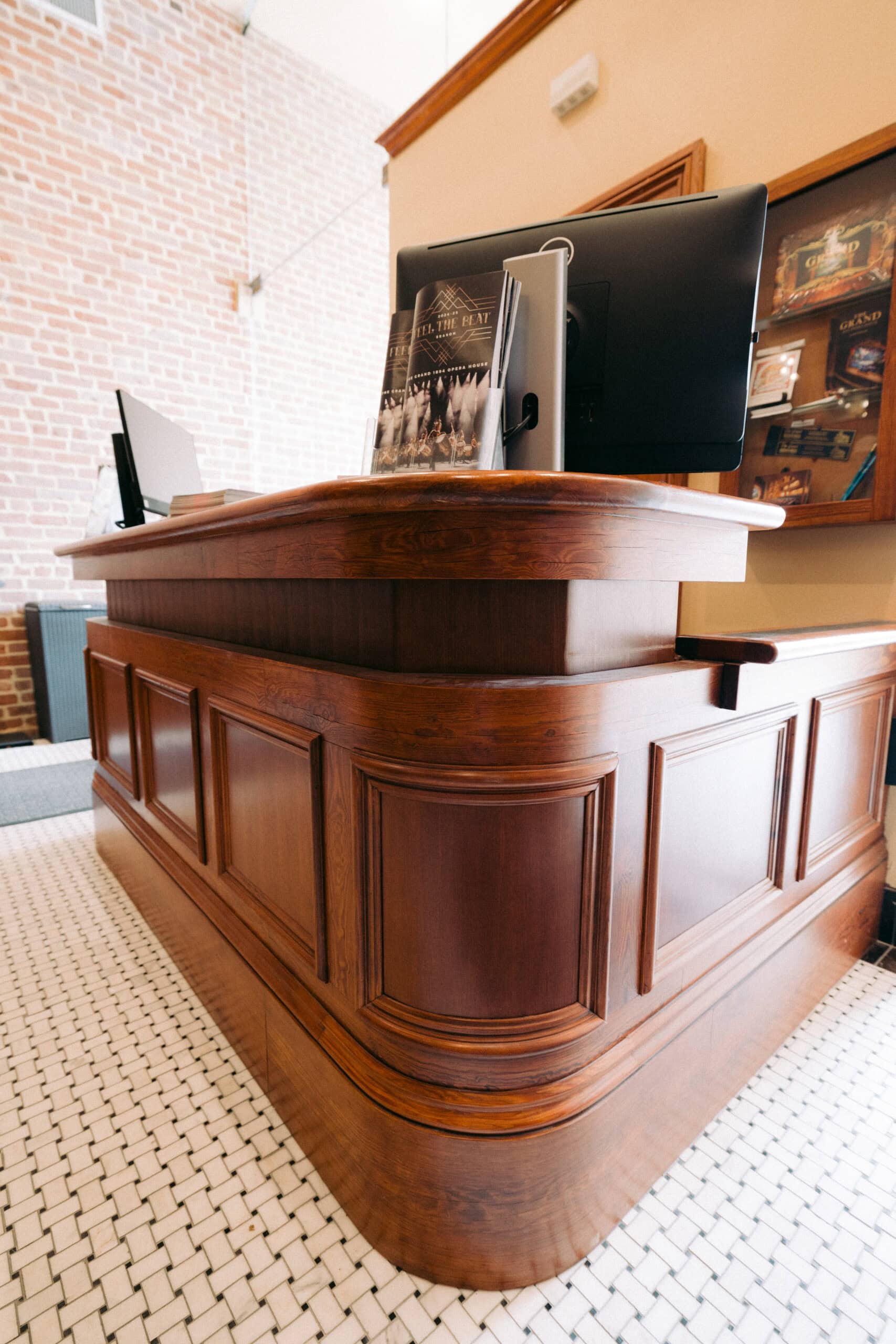 A wooden reception desk with paneled sides and two computer monitors welcomes guests to the Grand 1894 Opera House. A book or brochure is displayed on the desk, set against a brick wall, bookshelf, and white tile floor with a black pattern. - McNatt Contracting