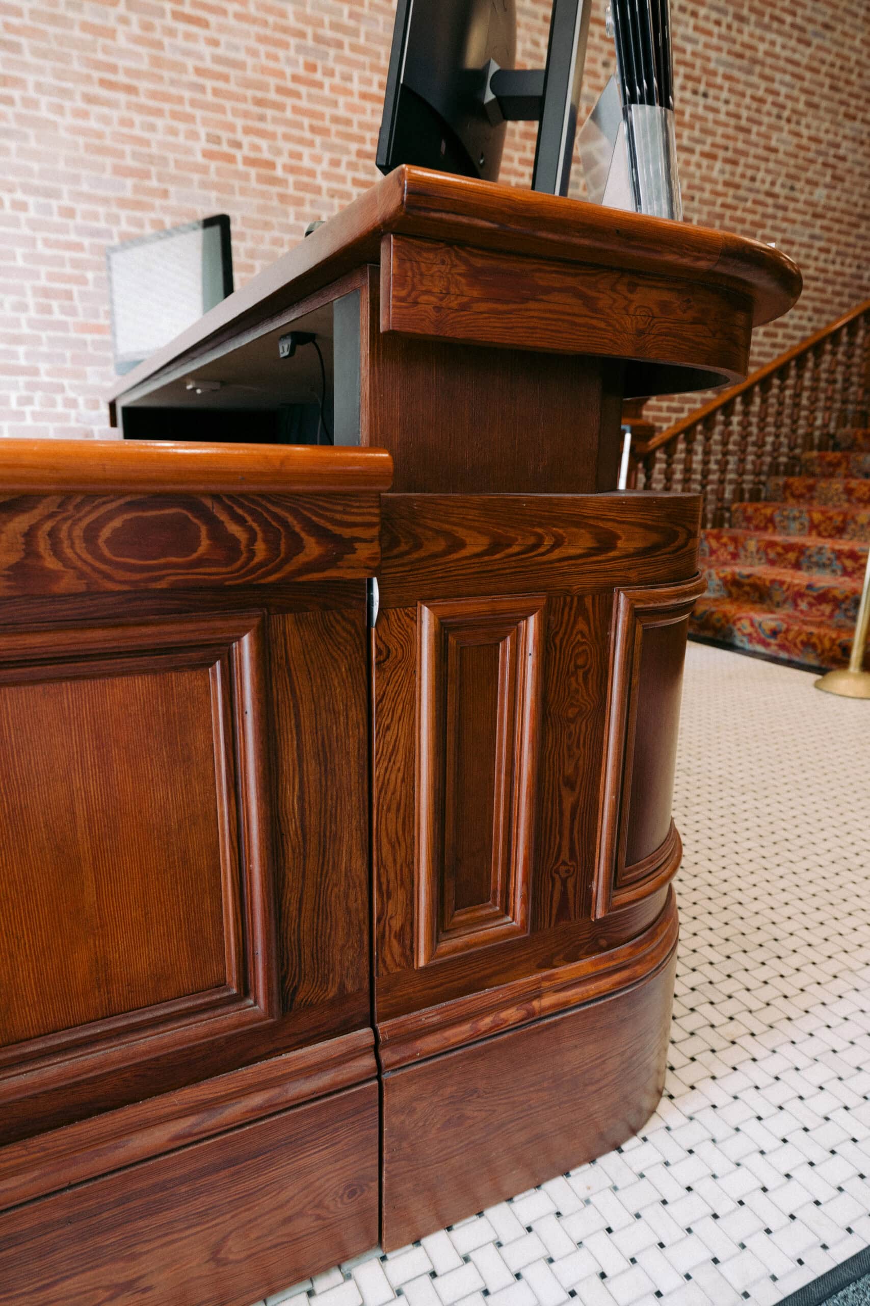 A close-up of a polished wooden reception desk with paneled detailing, set on a white tiled floor. A computer monitor and organizing trays are visible on the desk. A carpeted staircase and brick wall appear in the background. - McNatt Contracting