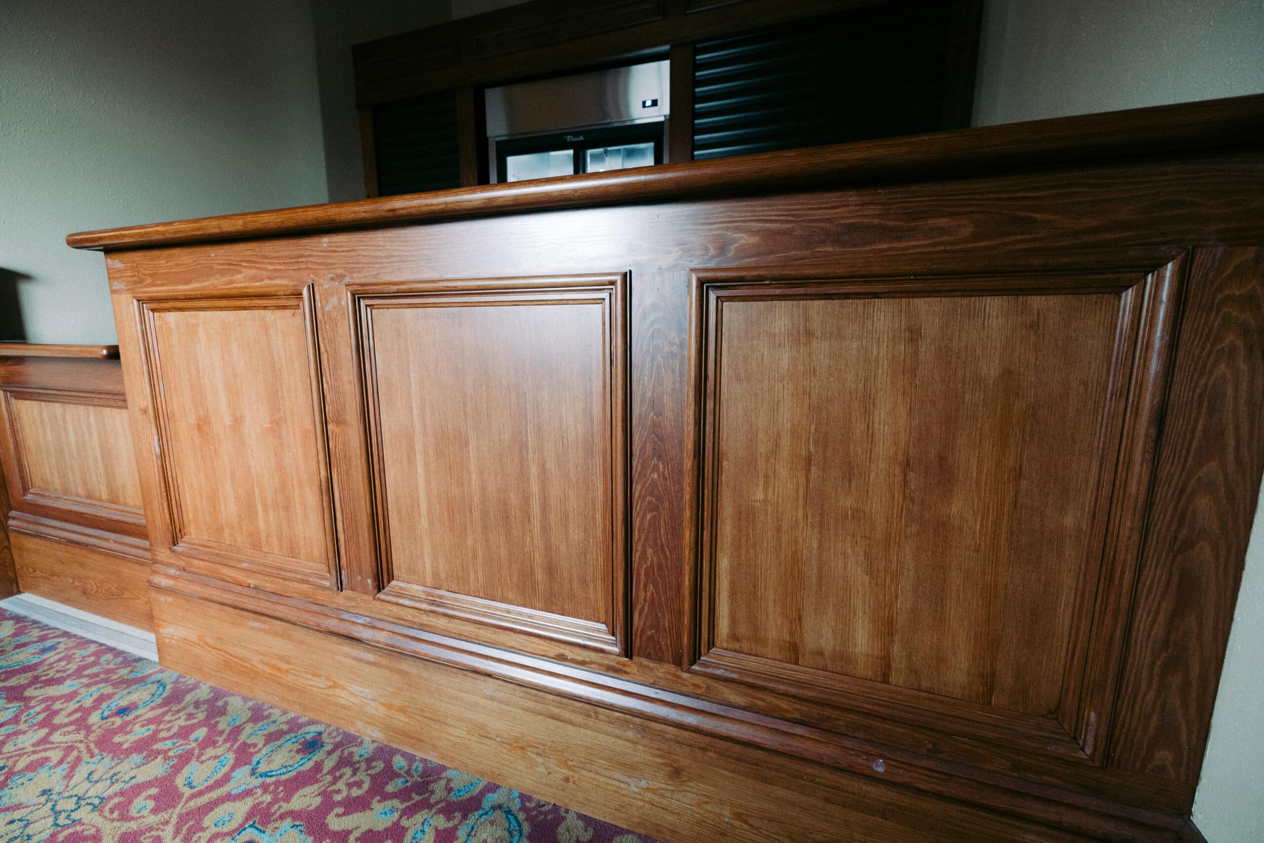 A wooden reception desk with rectangular panel details stands on a patterned carpet in front of a wall and a closed window or service hatch. - McNatt Contracting