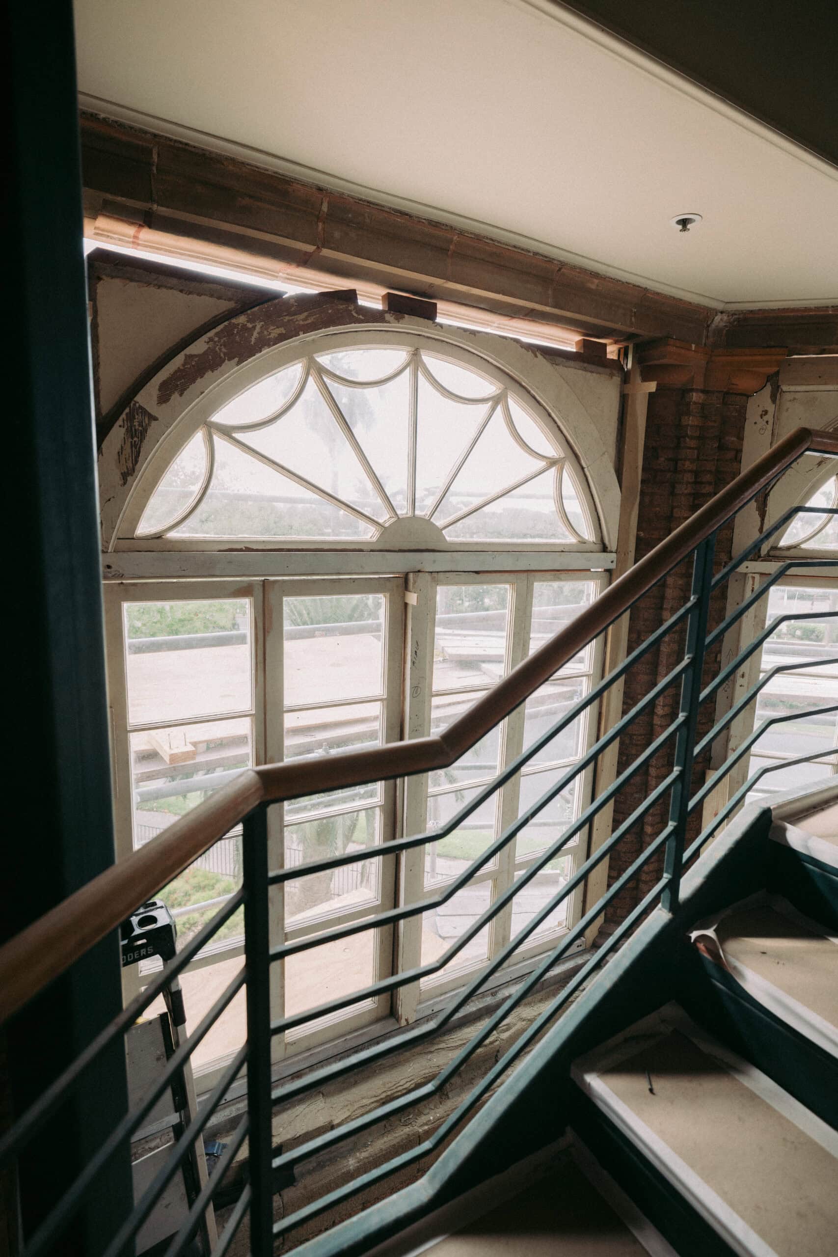 A staircase with a metal railing leads up next to a large, arched window with decorative panes. Sunlight comes through the window, which appears partially unfinished with exposed sections above. - McNatt Contracting