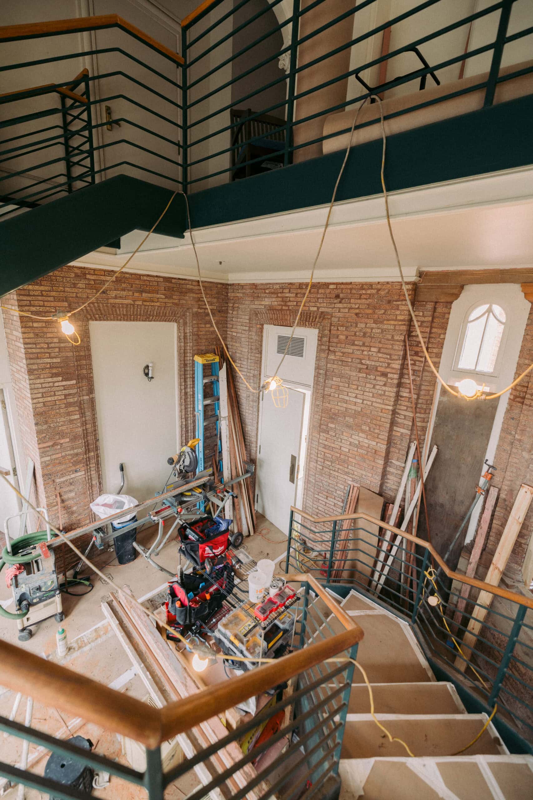 An indoor staircase surrounded by construction materials and tools, with exposed brick walls and hanging work lights, indicating ongoing renovation work in a multi-story building. - McNatt Contracting