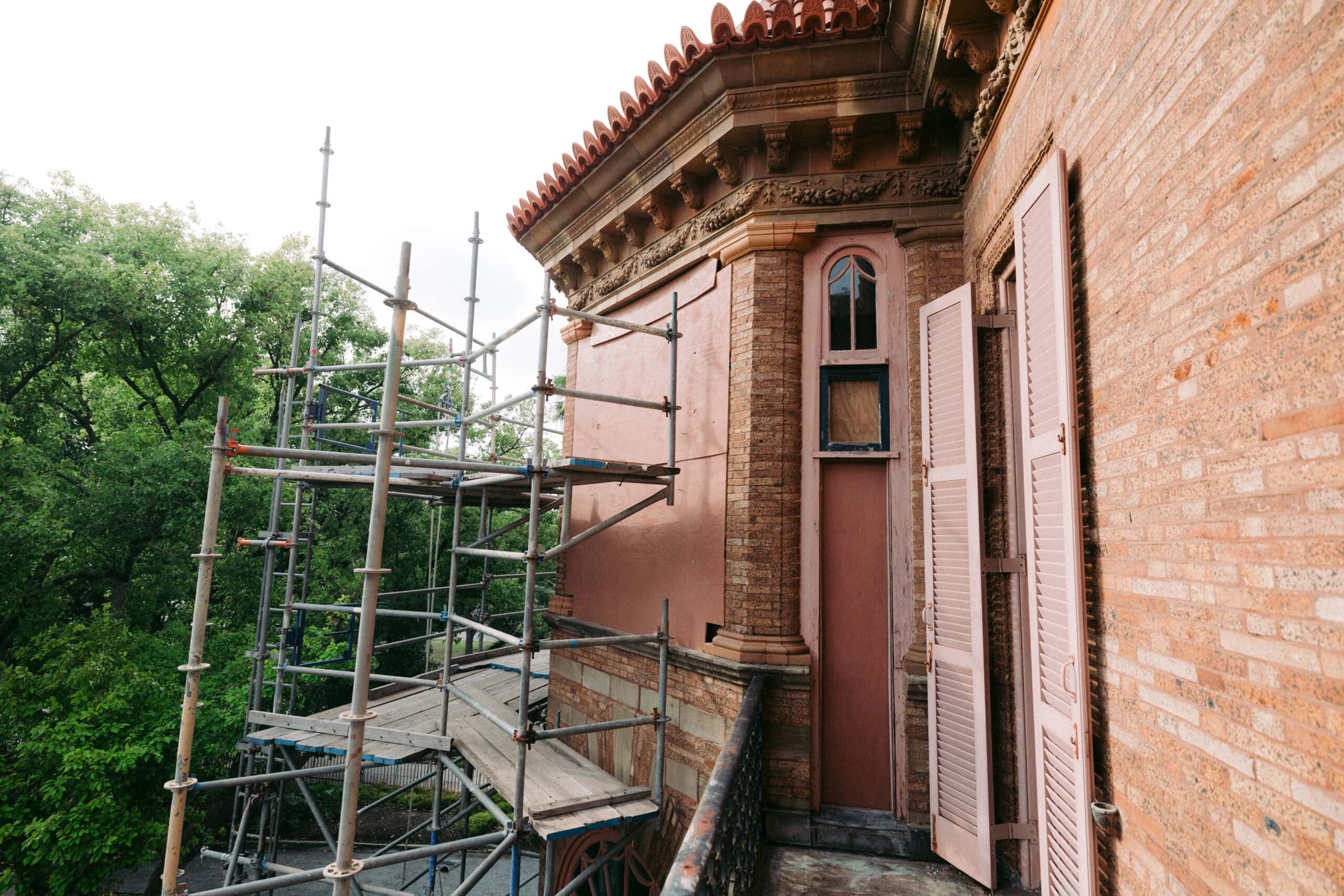 Scaffolding is set up next to an old brick building with pink shutters and ornate architectural details. Trees are visible in the background. - McNatt Contracting