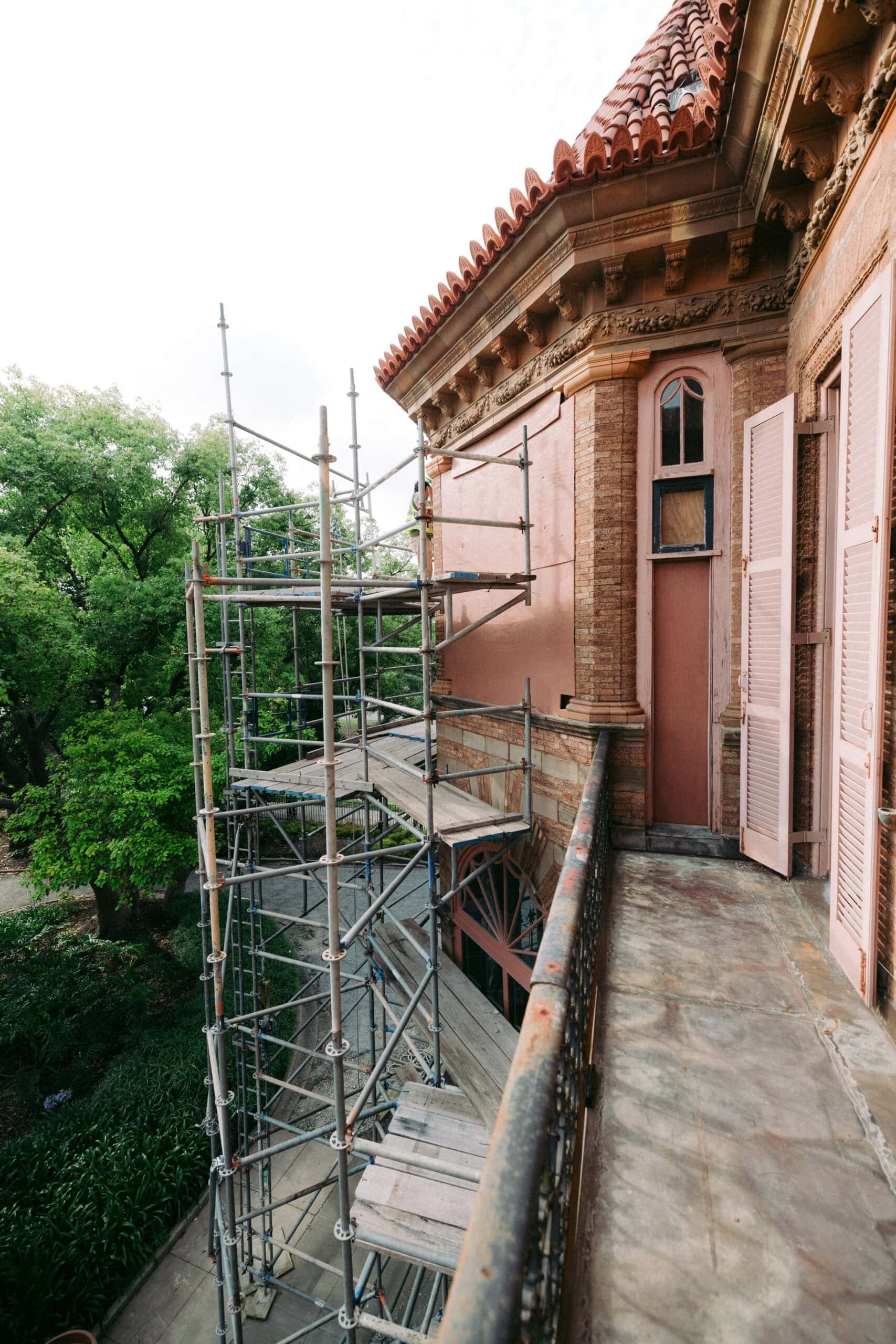 Scaffolding is set up alongside the exterior of a brick building with pink shutters and an ornate roof. The photo is taken from a balcony, with trees visible in the background. - McNatt Contracting
