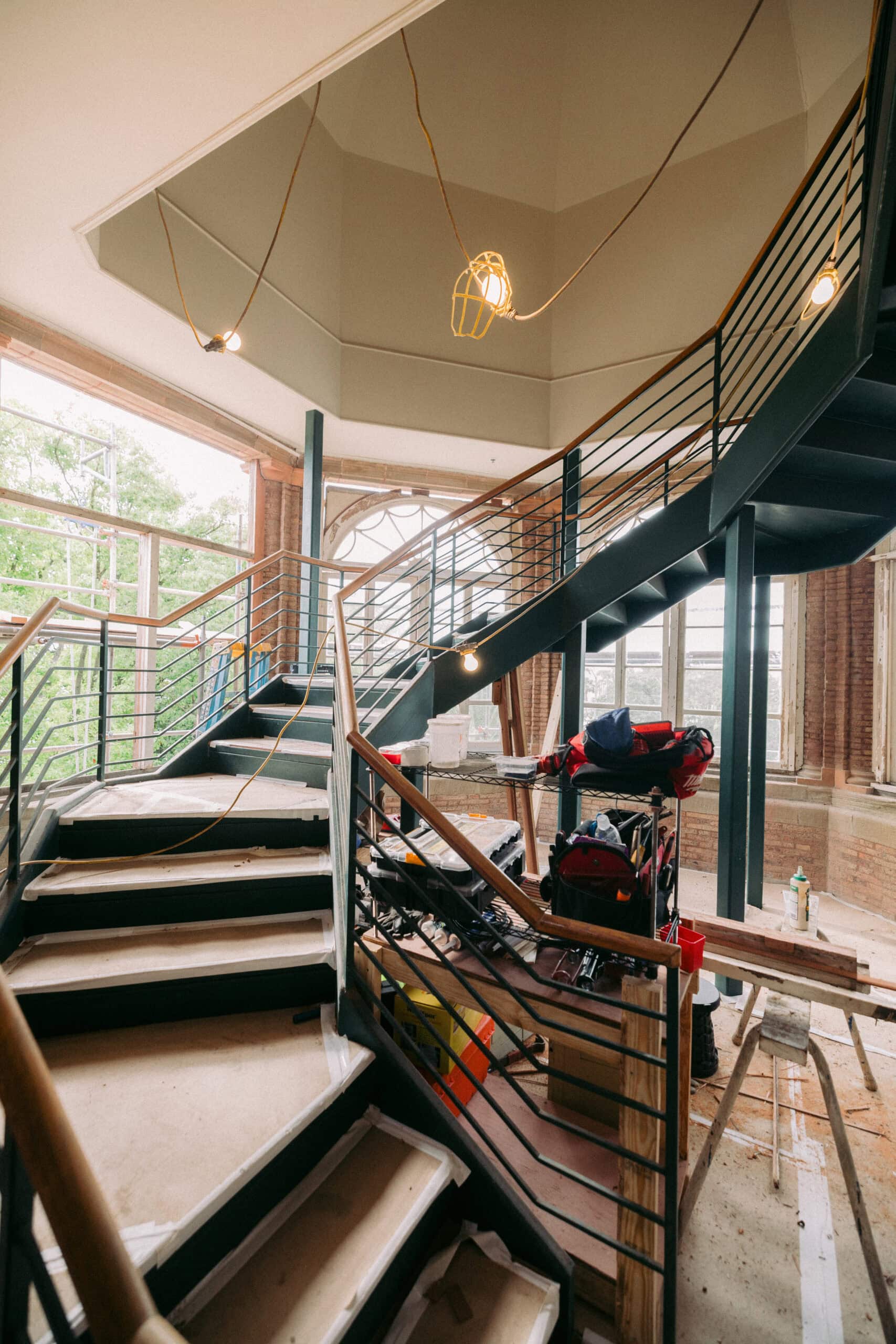 A modern metal staircase with wooden treads is under construction in a building with large windows. Construction tools, materials, and hanging work lights are visible throughout the area. - McNatt Contracting