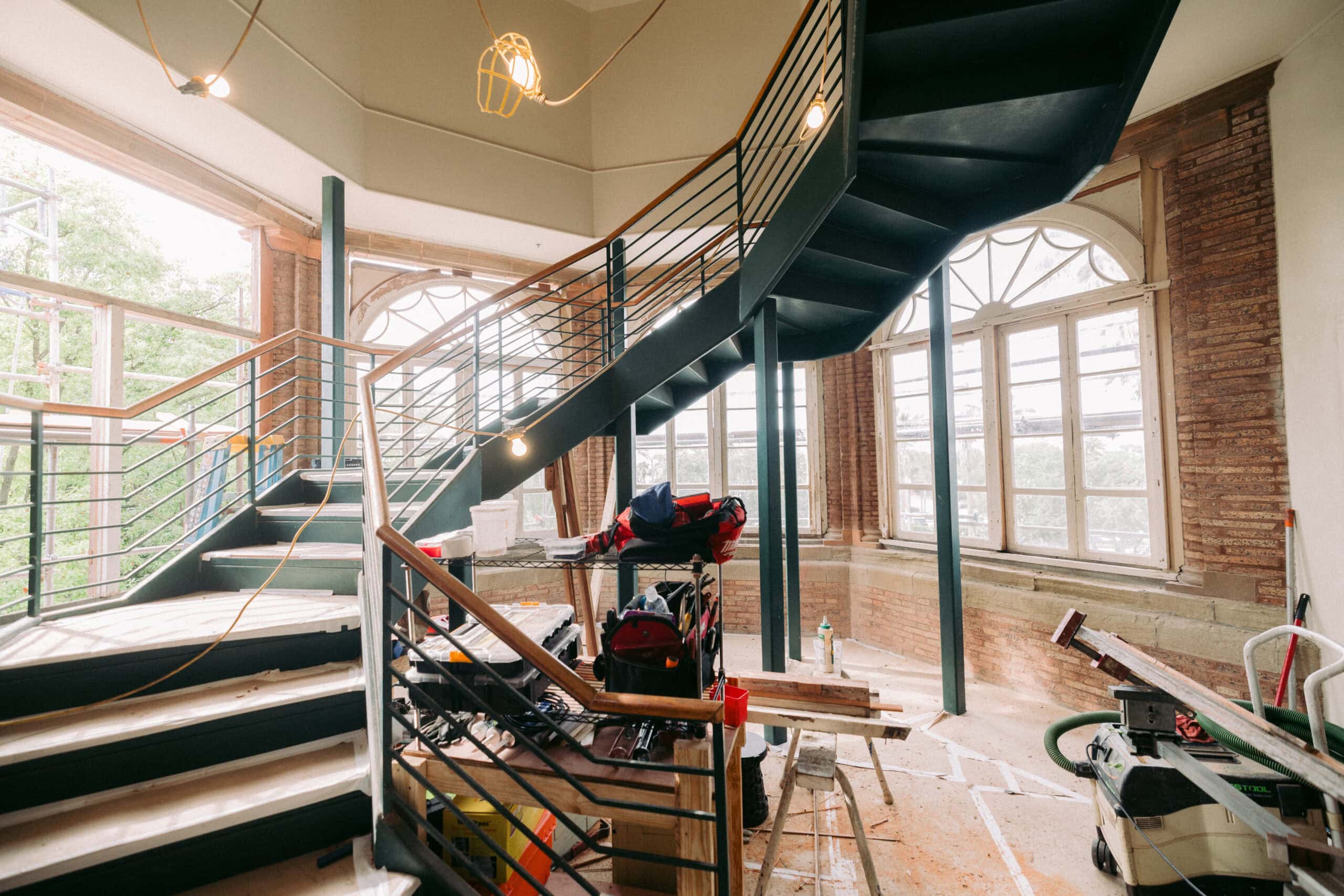 A spiral staircase under construction in a room with large windows, exposed brick walls, and various construction tools and materials scattered on the floor. - McNatt Contracting