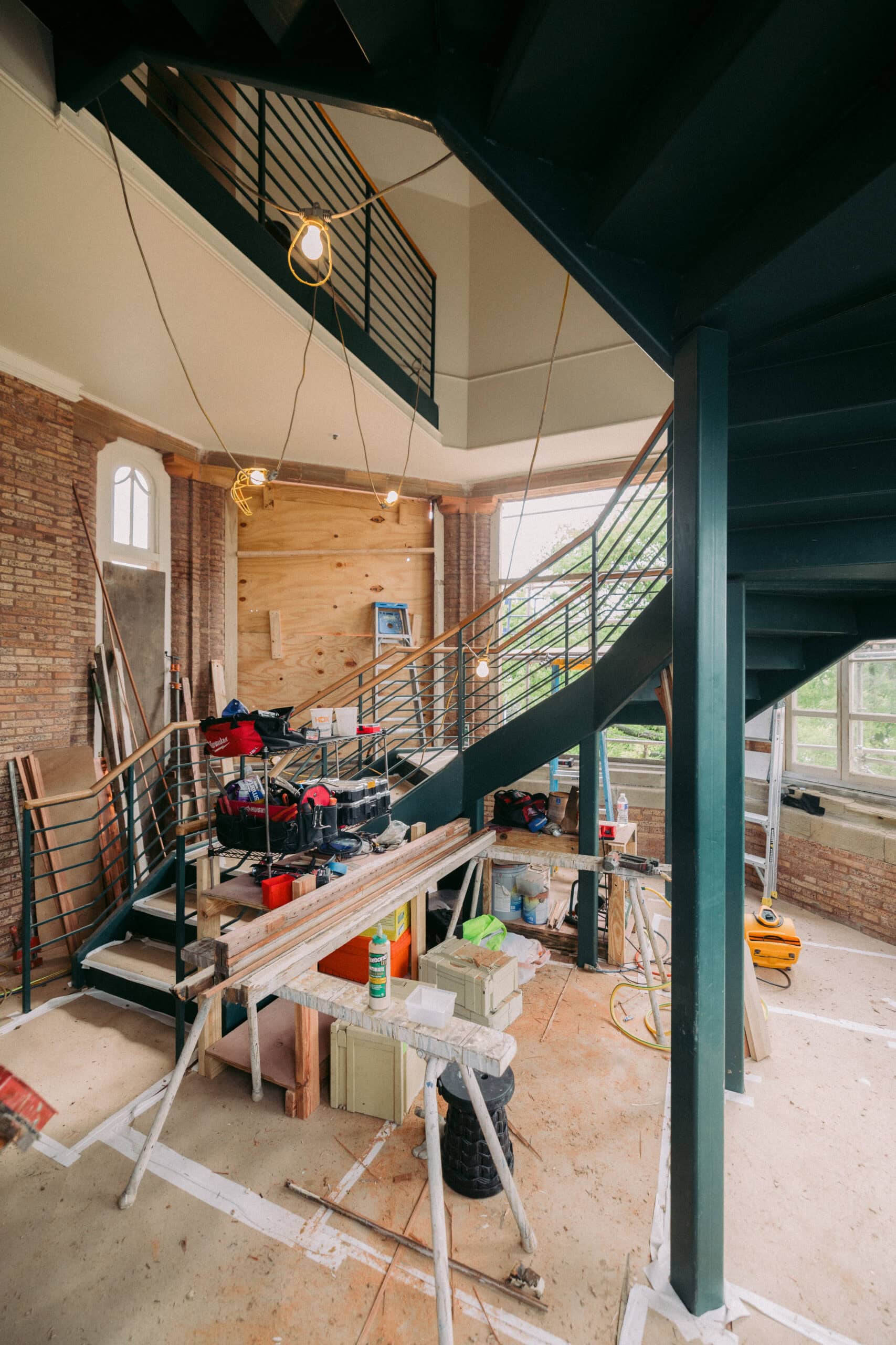 A modern staircase under construction in a room with exposed brick walls, scattered tools, workbenches, construction materials, and hanging light bulbs, indicating ongoing renovation work. - McNatt Contracting