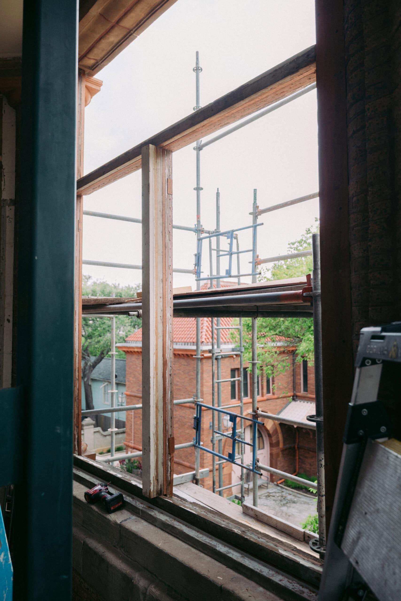 A window frame without glass overlooks scaffolding and a brick building with a tiled roof. Tools are visible on the windowsill and part of a ladder is on the right side of the image. - McNatt Contracting