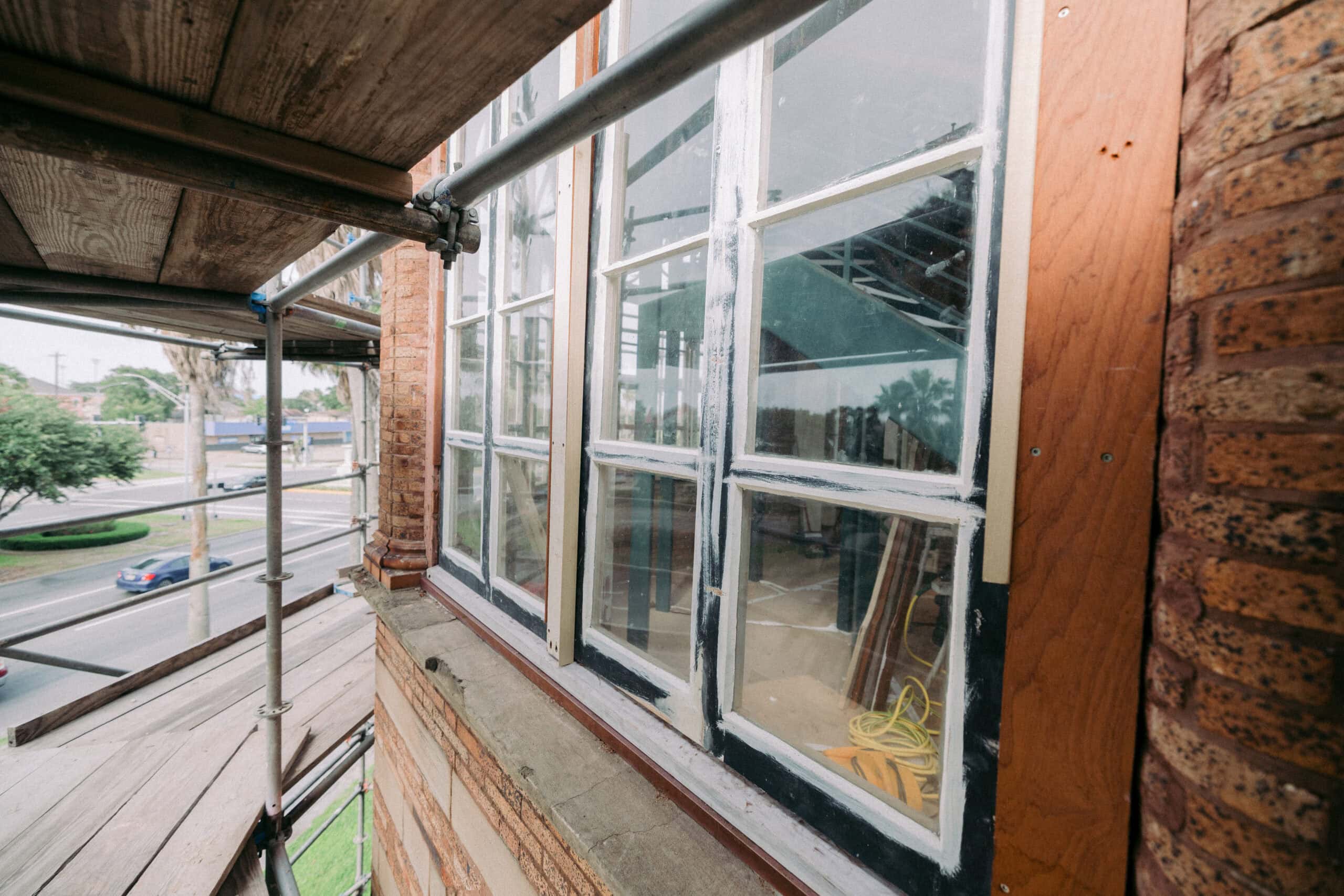 Scaffolding is set up along the exterior of a brick building under construction or renovation; new window frames are visible, and construction tools can be seen inside through the glass. - McNatt Contracting