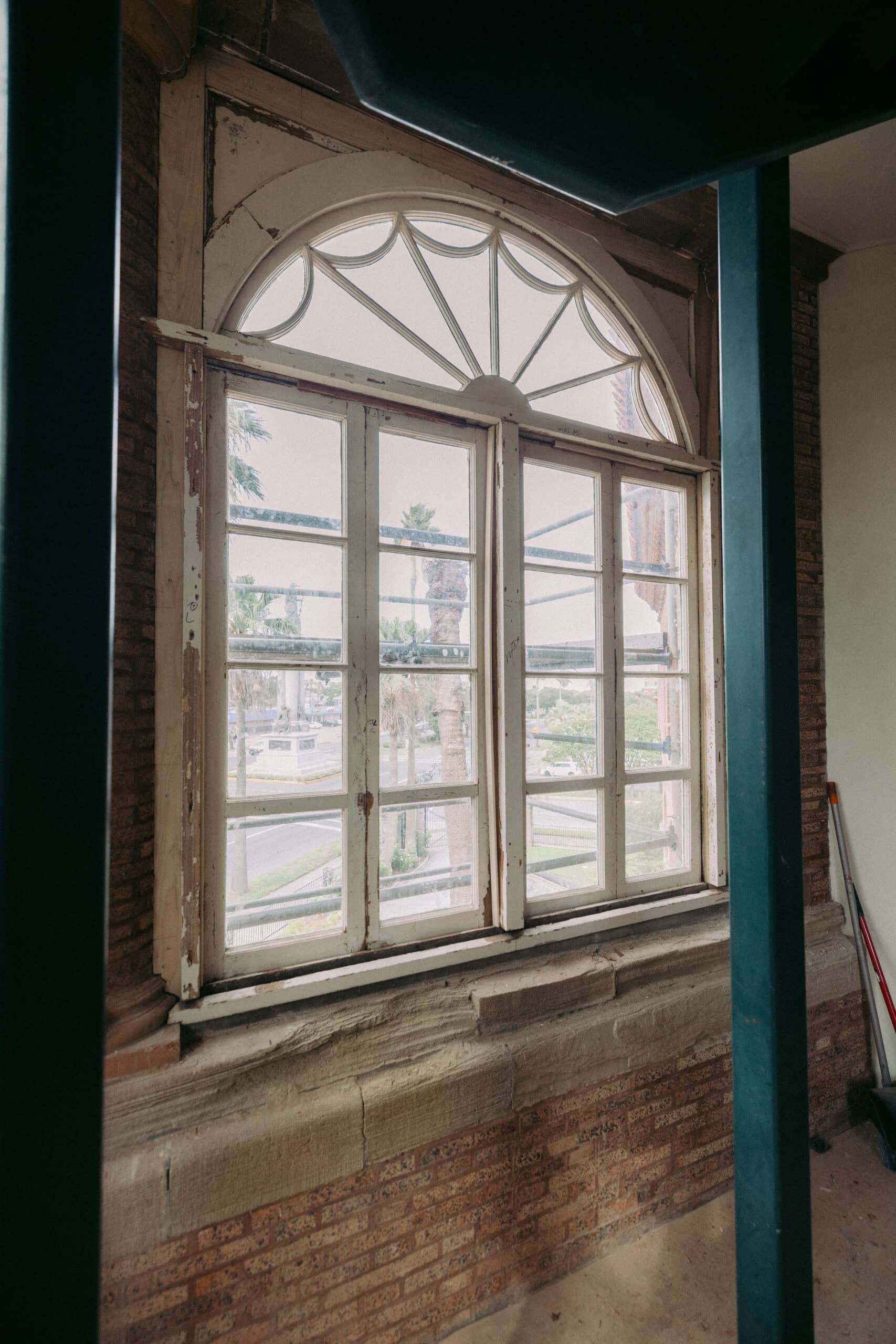 An old arched window with peeling white paint, set in a brick wall. The window overlooks a street with palm trees visible outside. The interior shows some exposed beams and a broom in the corner. - McNatt Contracting