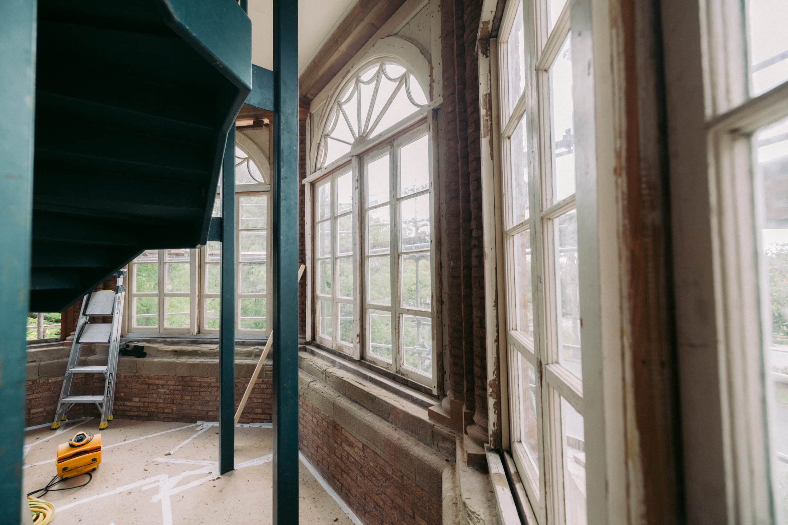 A room under renovation with large windows, a spiral staircase, a ladder, and a yellow vacuum on the floor. Sunlight is coming through the windows, and the walls have exposed brick and plaster. - McNatt Contracting