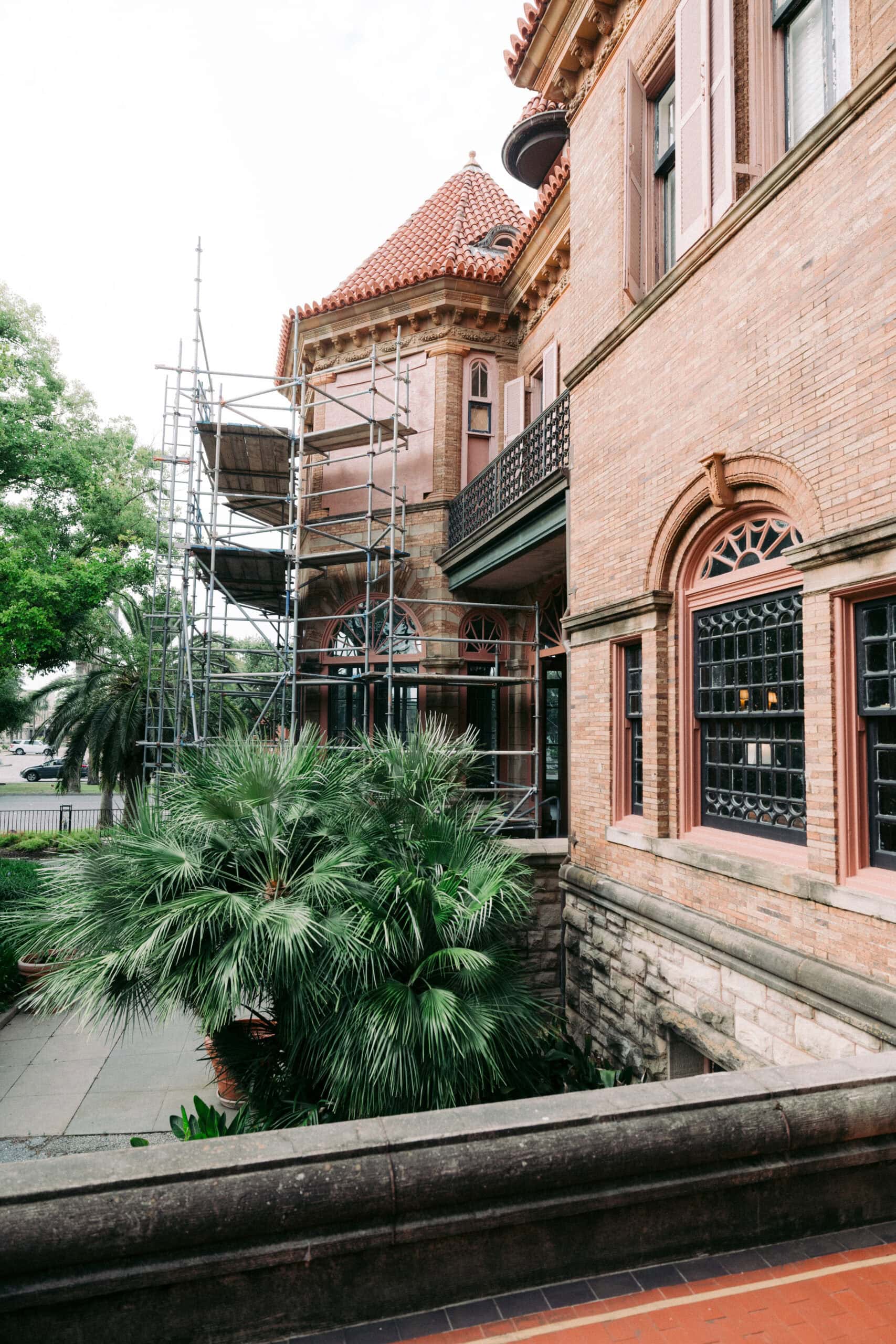Scaffolding is set up along the side of a historic brick building with arched windows and a red tile roof. Palm trees and greenery are visible in the foreground. - McNatt Contracting
