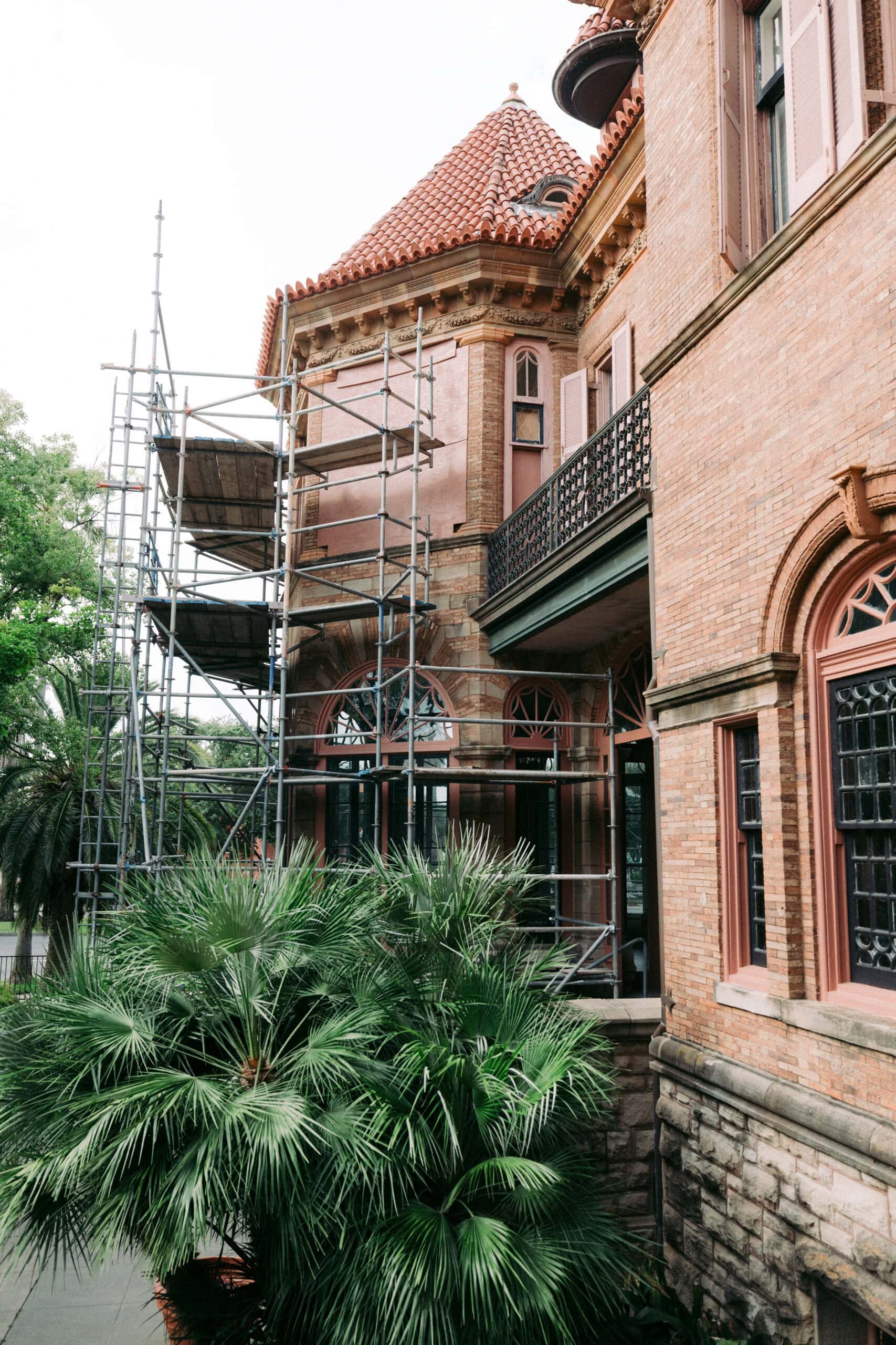 A brick building with arched windows and a red-tiled roof has scaffolding set up along one side. There are green palm trees in the foreground and some overcast lighting. - McNatt Contracting