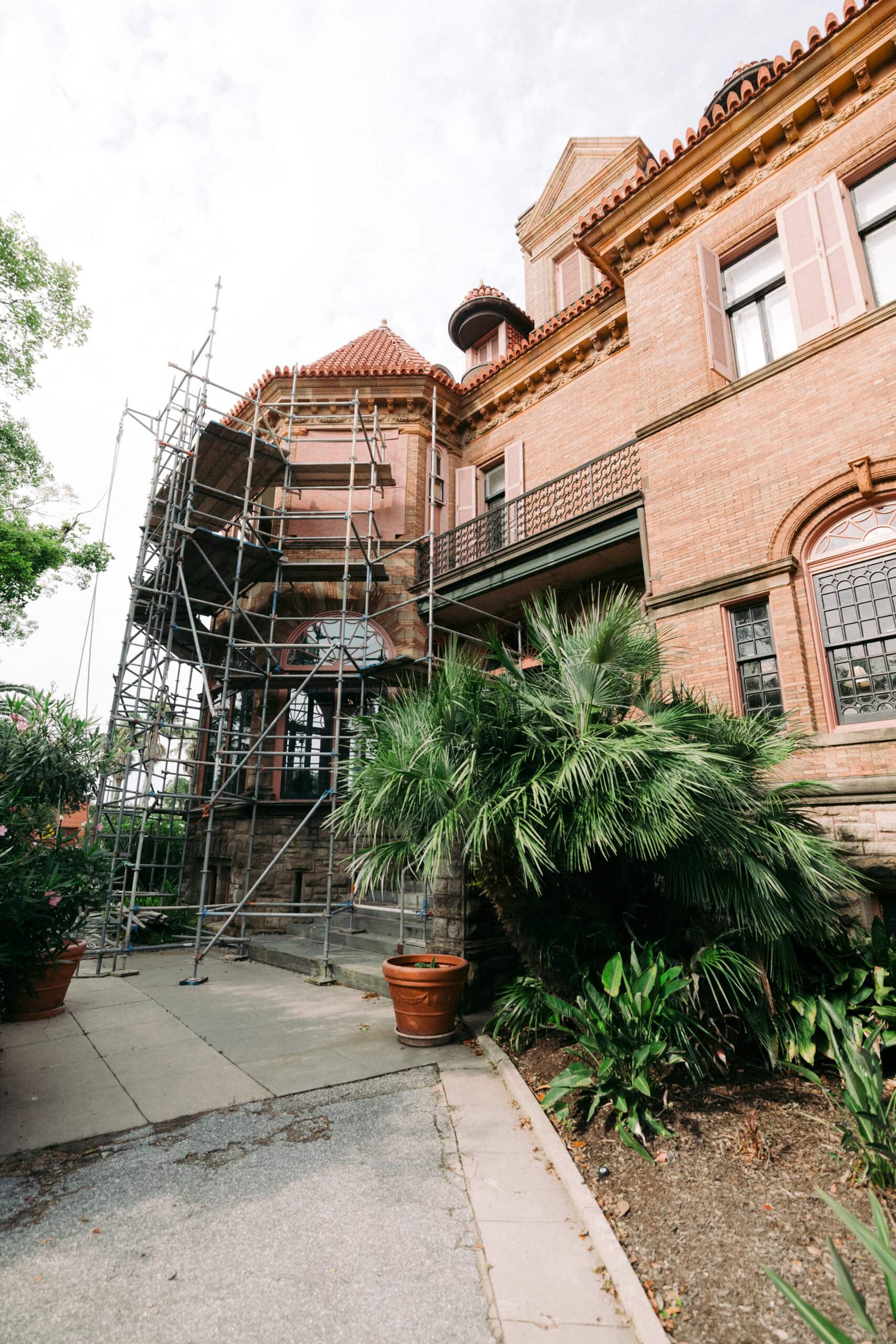 A large brick building with arched windows and a tile roof has metal scaffolding set up along one side. Lush green plants and potted palms line the walkway in front of the building. - McNatt Contracting
