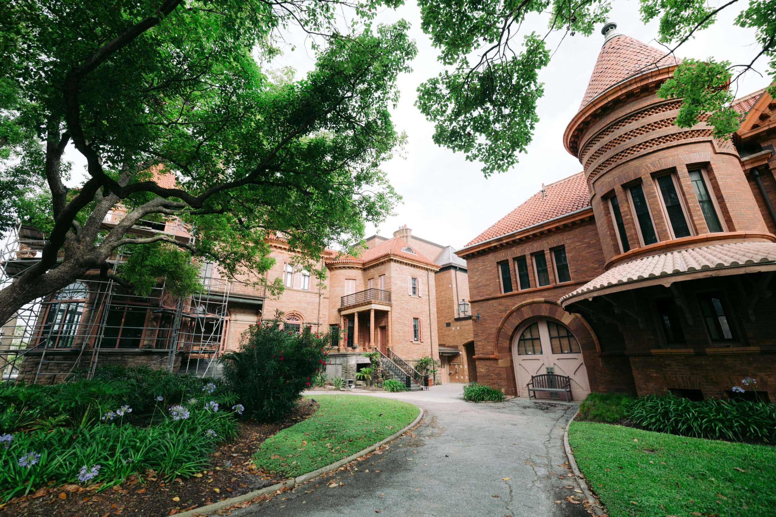 A large, historic brick building with turrets and arched windows, surrounded by green trees and gardens. Scaffolding is visible on the left side of the building. A curved driveway leads to the entrance. - McNatt Contracting