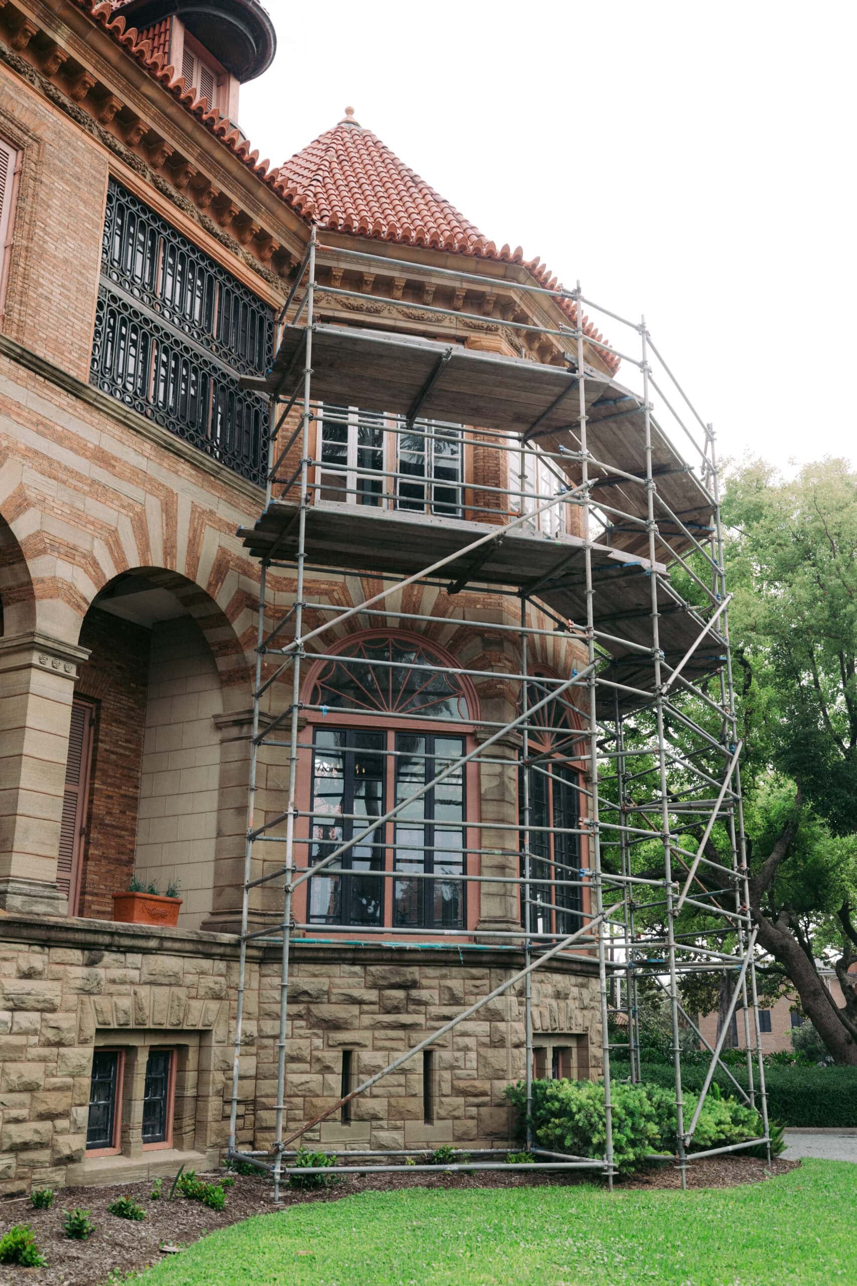 Scaffolding is set up against the exterior of a historic brick and stone building with arched windows and a red tiled roof. Green grass and trees are visible in the foreground. - McNatt Contracting