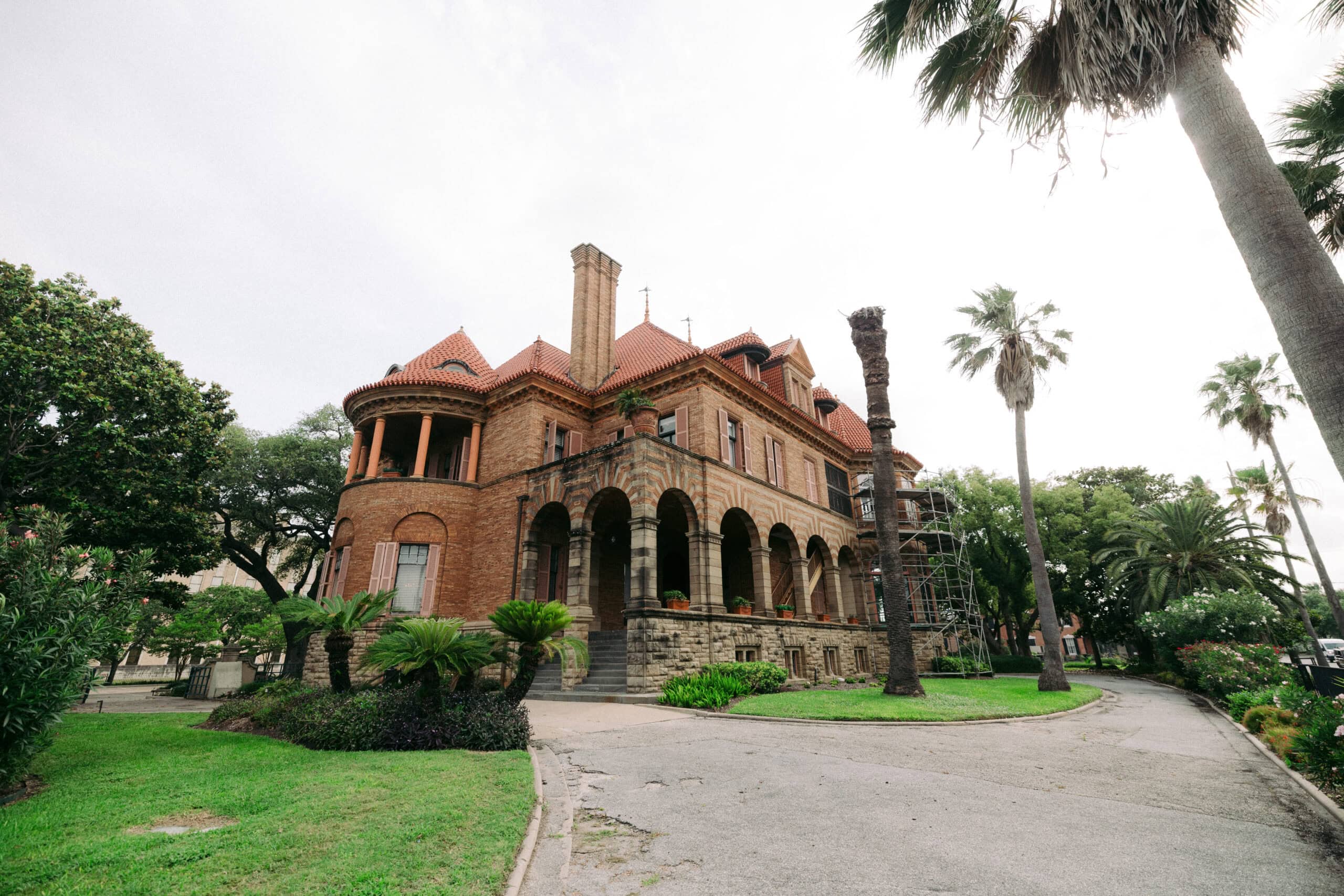 The Sealy Mansion, also known as Open Gates, is a large historic brick mansion with arched windows, multiple chimneys, and red-tiled roofs, nestled amid lush lawns, palm trees, and landscaped greenery on a sunny day. - McNatt Contracting