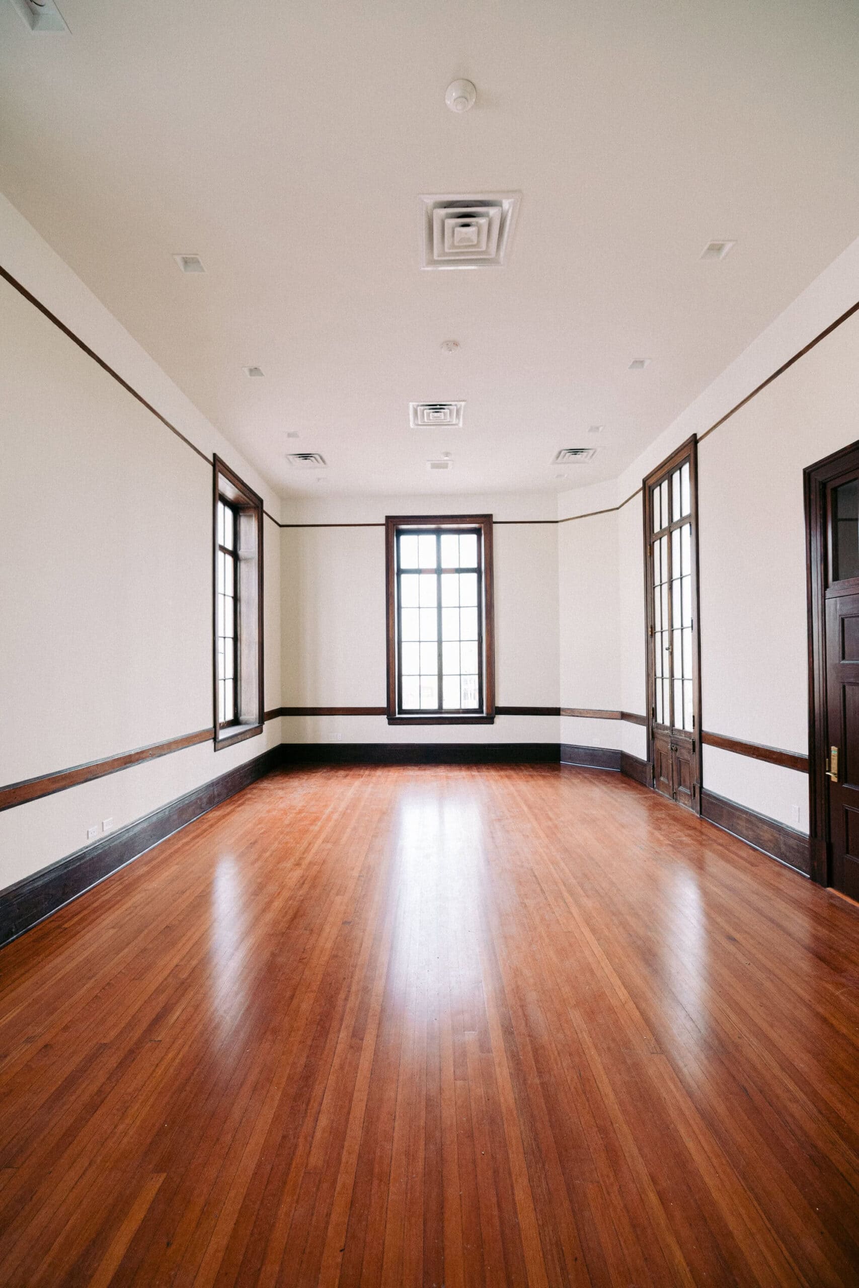 A spacious, empty room with wooden floors, white walls, large windows letting in natural light, and dark wood trim and doors. Ceiling features several air vents and recessed lights. - McNatt Contracting