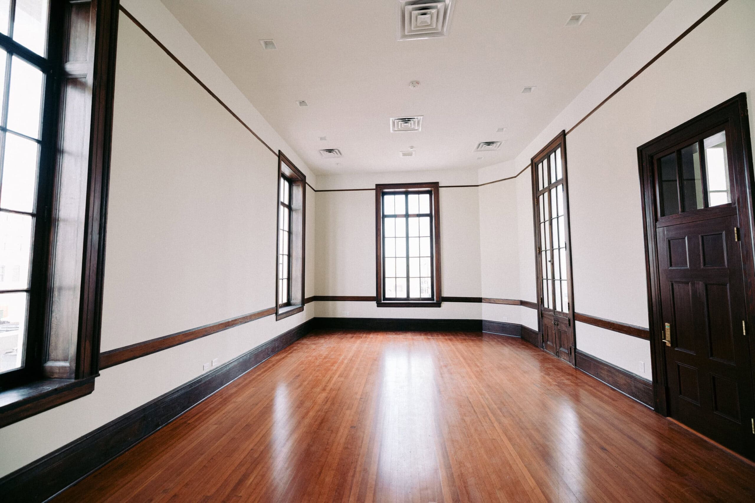 Empty room with polished wooden floors, cream-colored walls, tall windows with dark wood trim, and matching dark wood doors. Natural light fills the space from the large windows. - McNatt Contracting