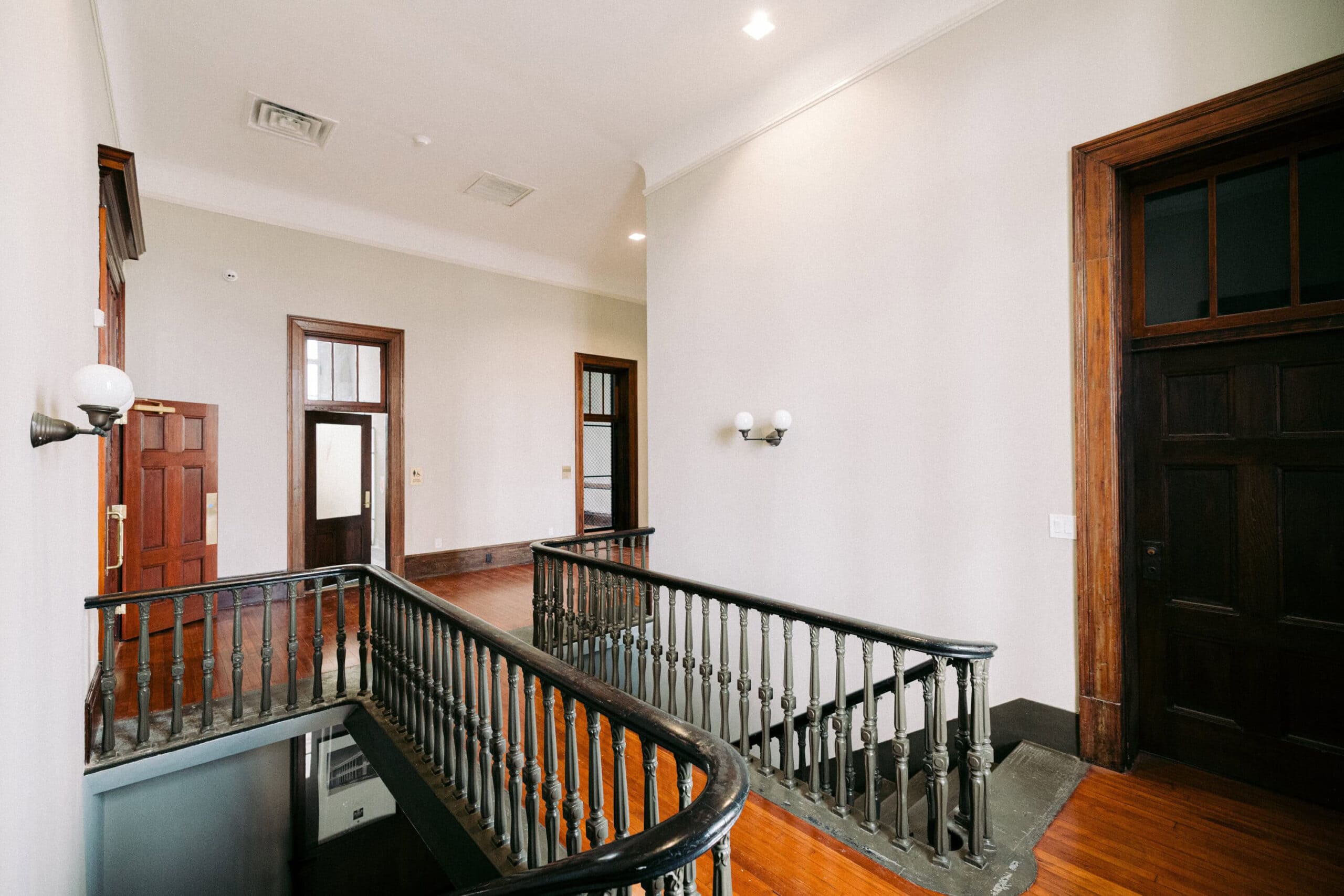 An indoor landing area with polished wooden floors, white walls, a black metal railing around a staircase, and several wooden doors leading to adjacent rooms. Wall-mounted lamps provide lighting. - McNatt Contracting