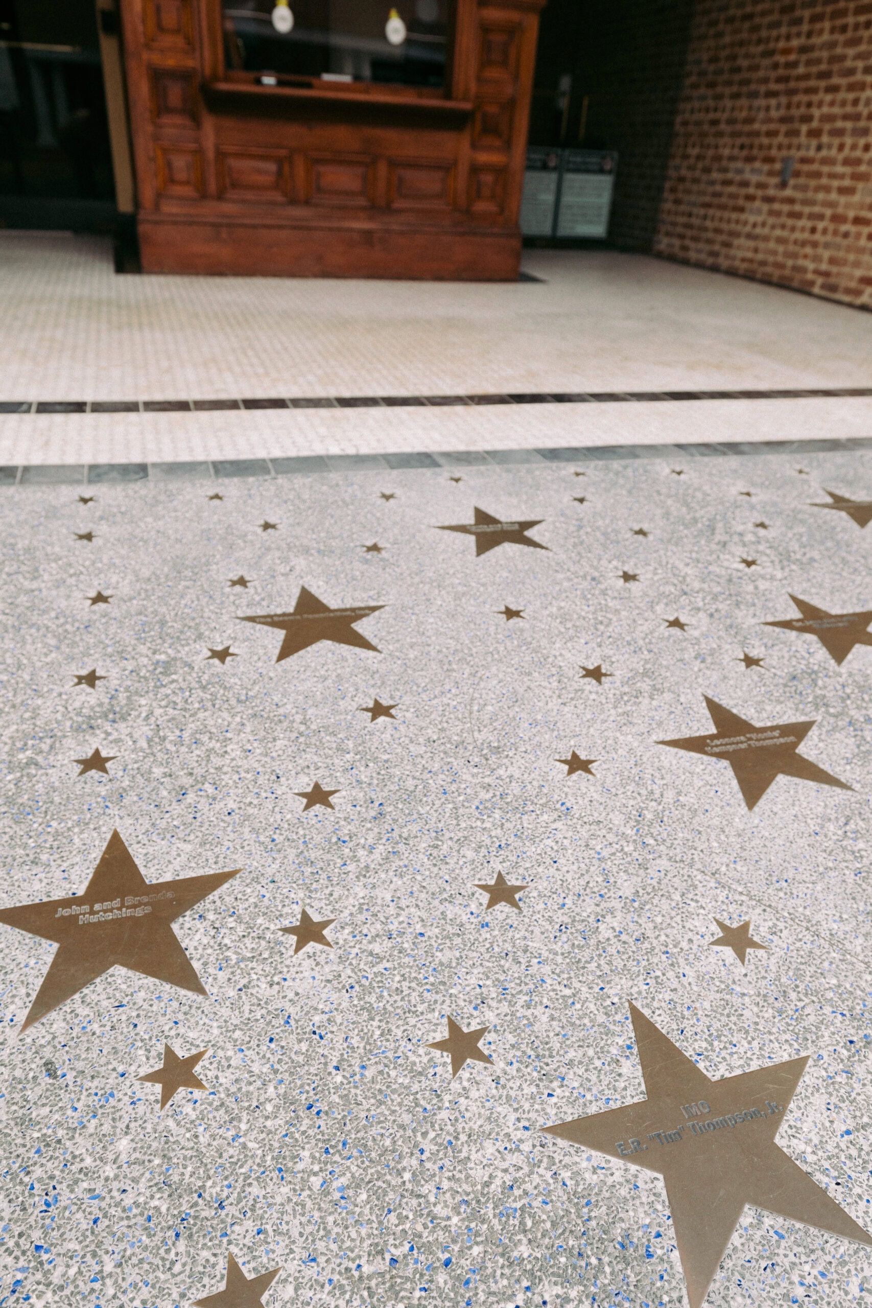 Bronze stars embedded in a speckled sidewalk lead up to a wooden building entrance with glass doors and a brick wall. Some stars contain engraved names, though the text is not clearly readable. - McNatt Contracting