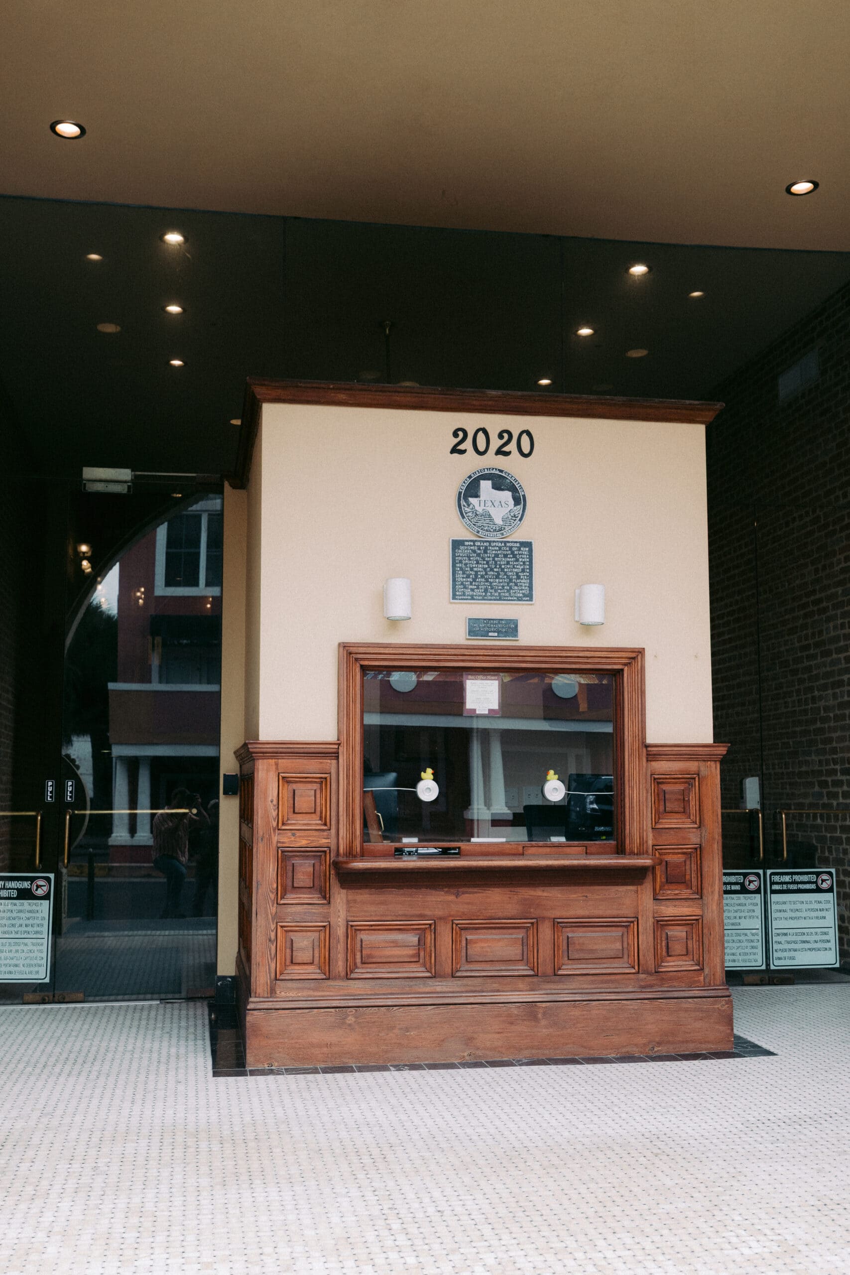 A wooden ticket booth with glass windows stands at the entrance of a building, hinting at the charm of Historic Theaters Galveston. The number 2020 and a circular plaque are displayed above it, while tiled flooring surrounds this inviting Opera House Galveston scene. - McNatt Contracting